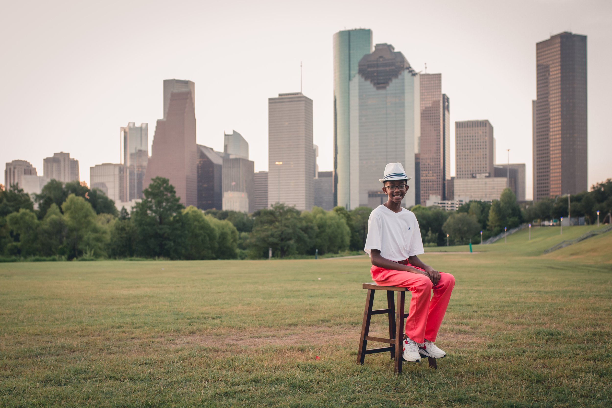 A smiling young man sitting on a stool in a grassy park with a city skyline in the background.