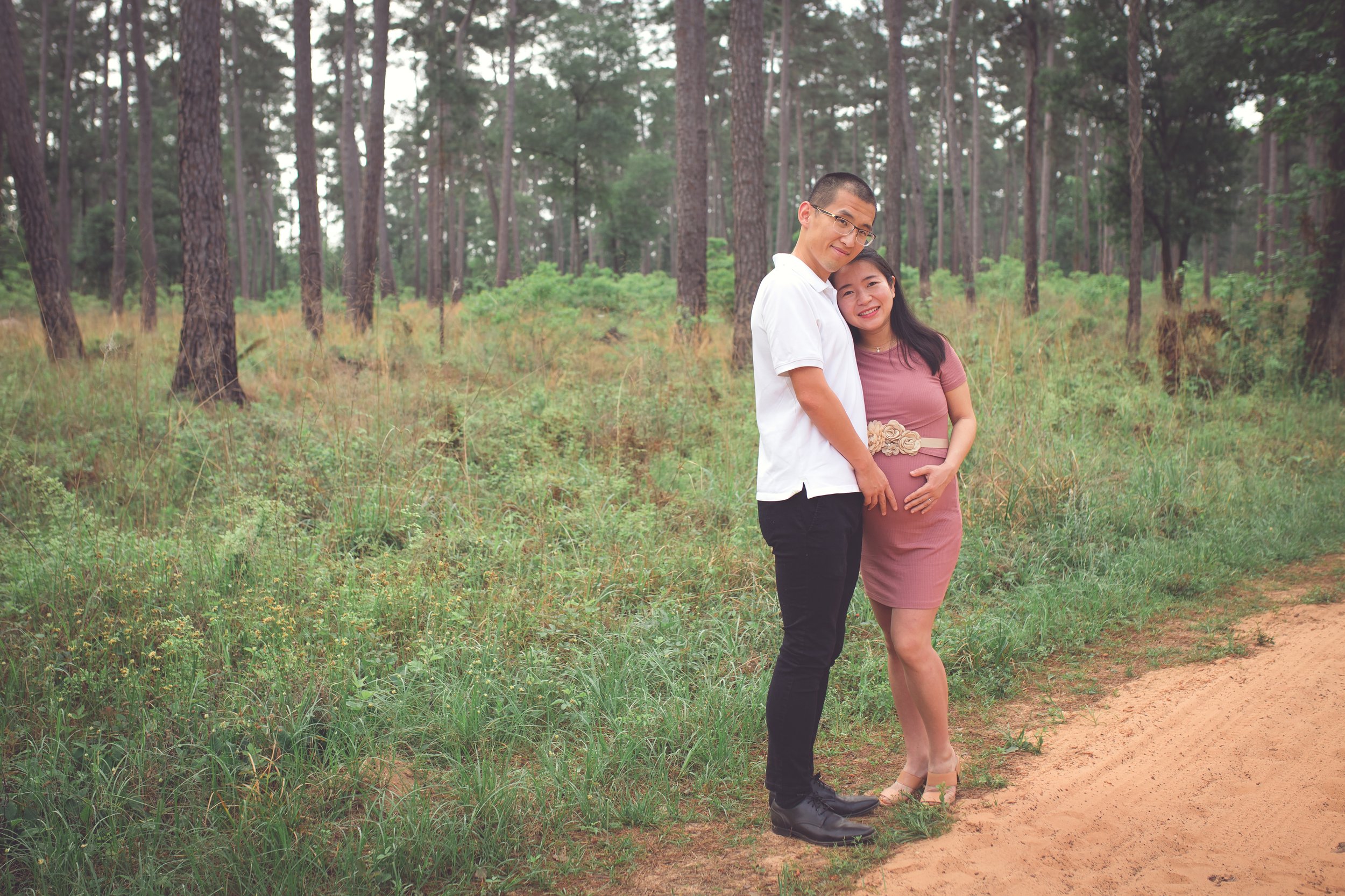 A couple standing in a forest, smiling, with the woman holding her pregnant belly, both dressed casually.