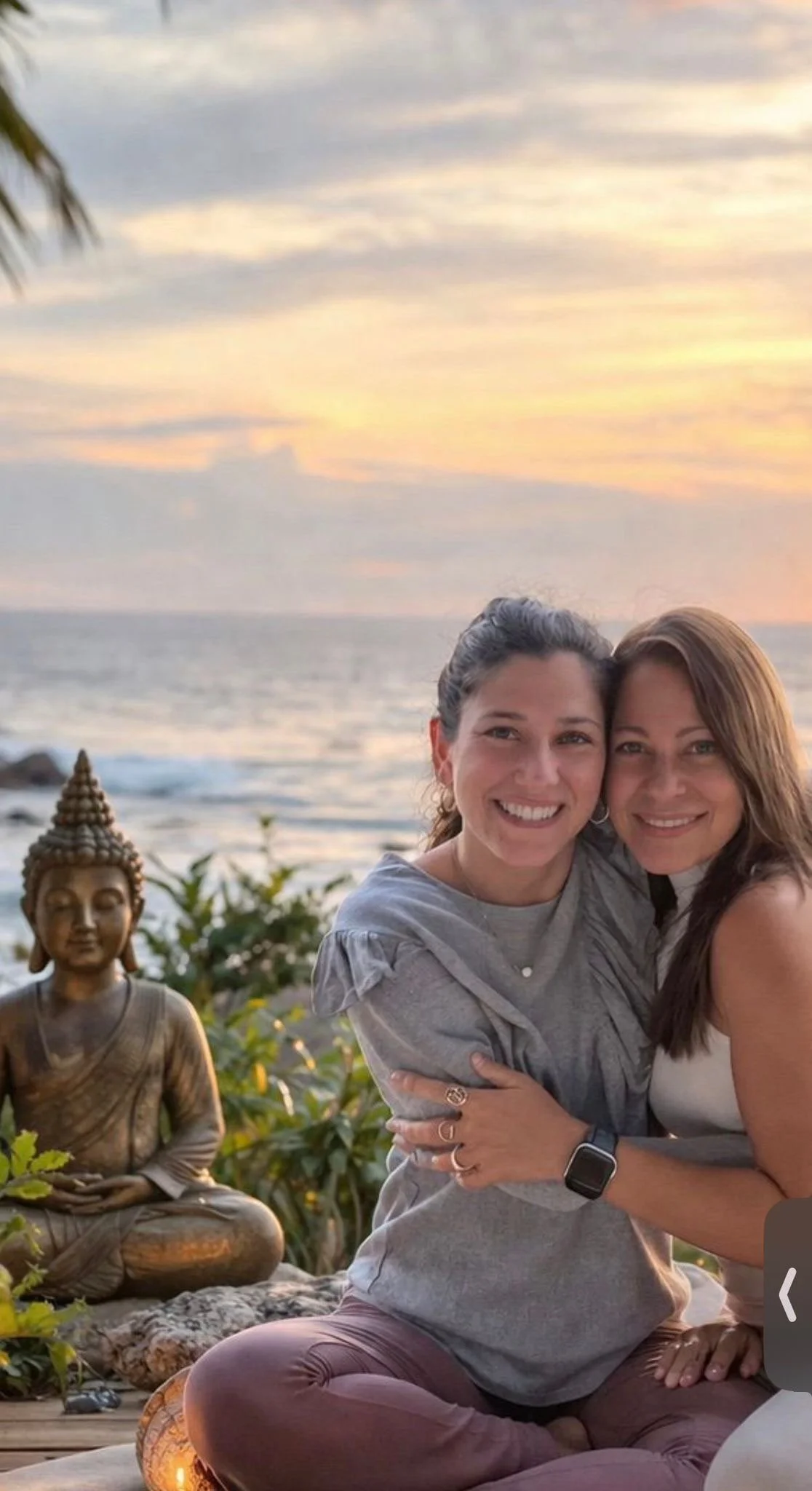 Two women with long hair, smiling and sitting closely together with their arms around each other, outdoors at sunset near the ocean, with a small Buddha statue and greenery nearby.