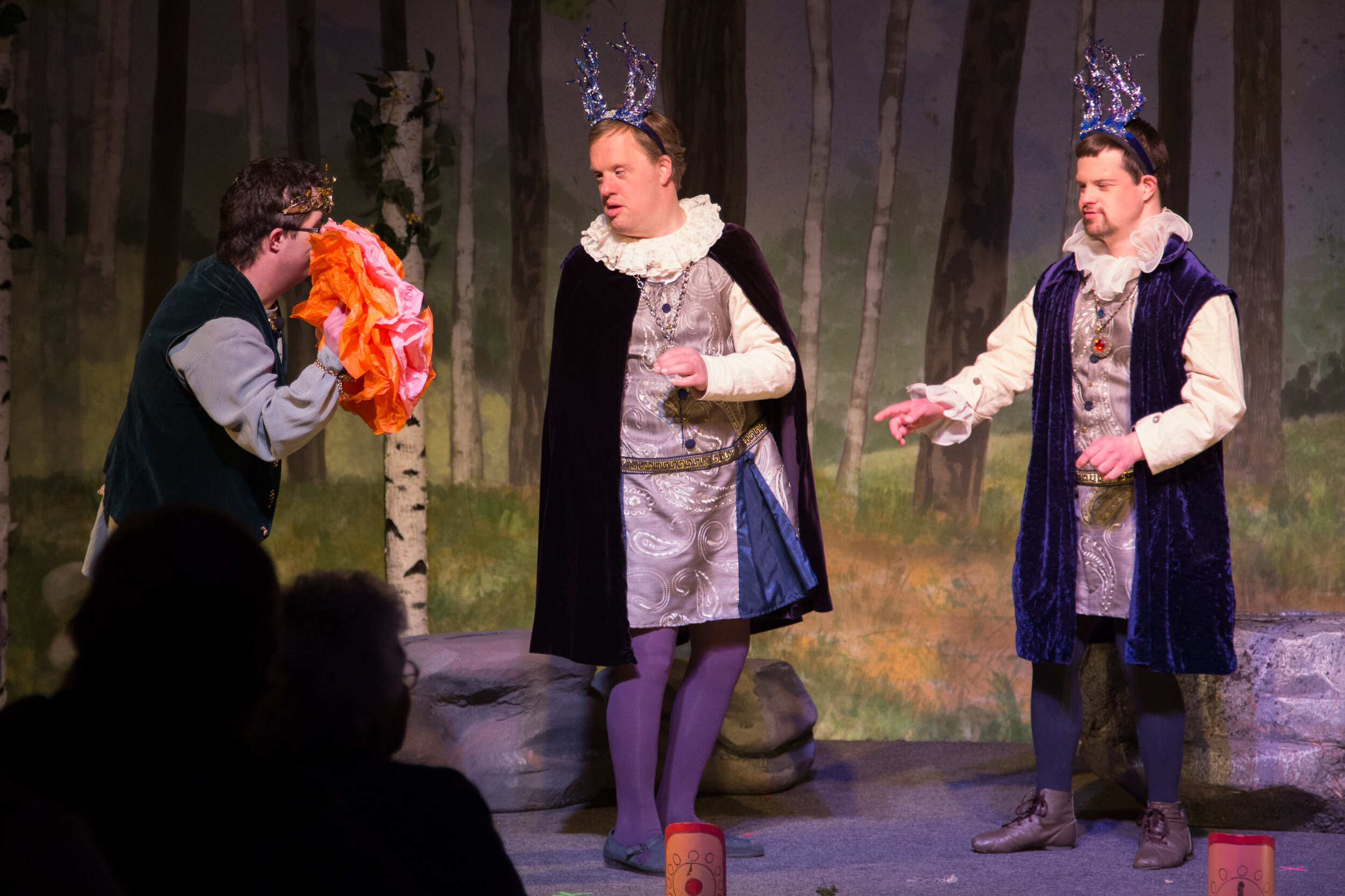 Three actors in decorative crowns and Shakespearean costumes perform on a stage with scenic rocks, a painted forest behind them. We can see some audience members in silhouette sitting in the foreground of the image.