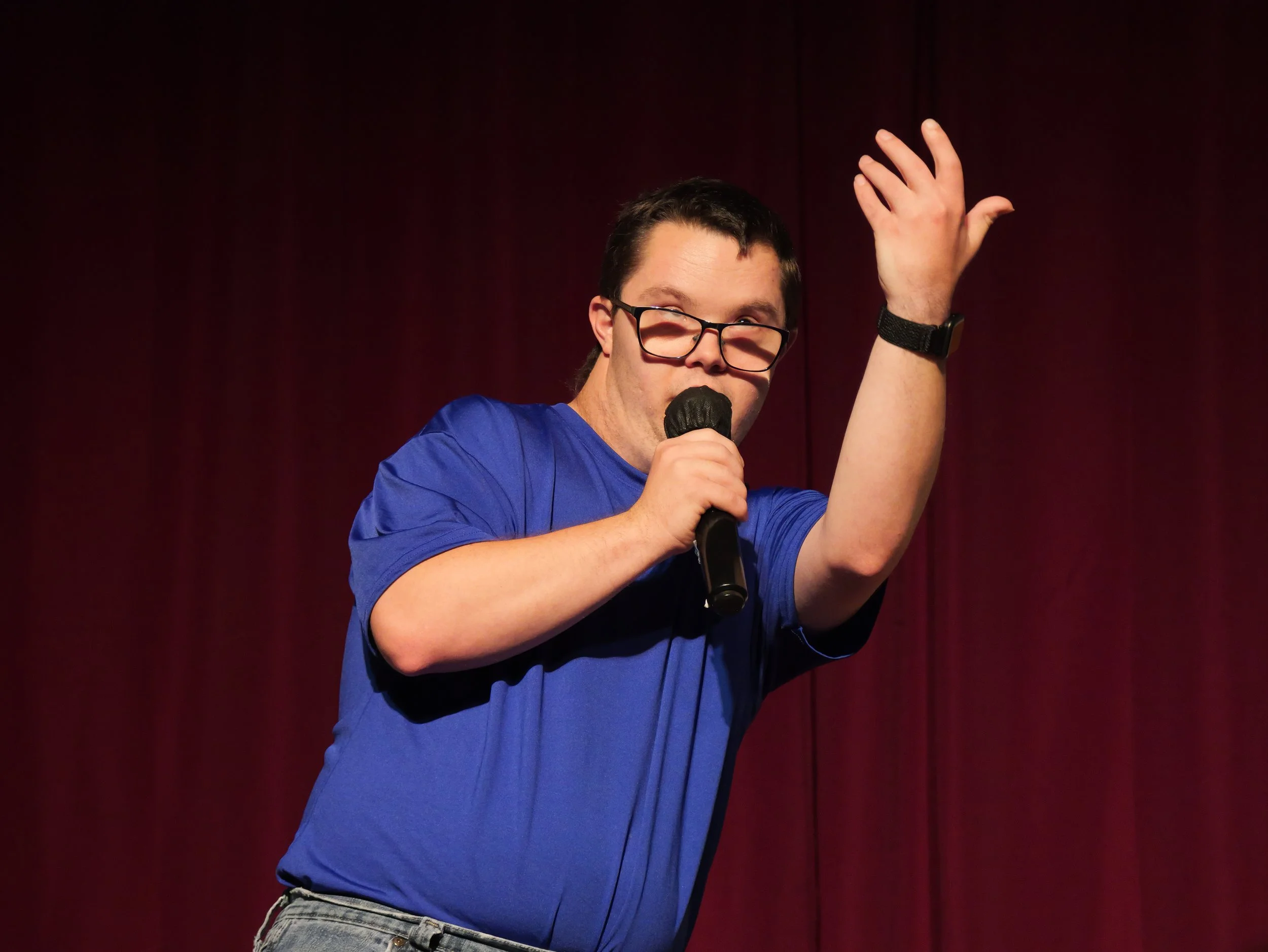 An actor wearing glasses, a blue T-shirt, and jeans sings into a handheld microphone while raising one hand expressively onstage in front of a dark red curtain.
