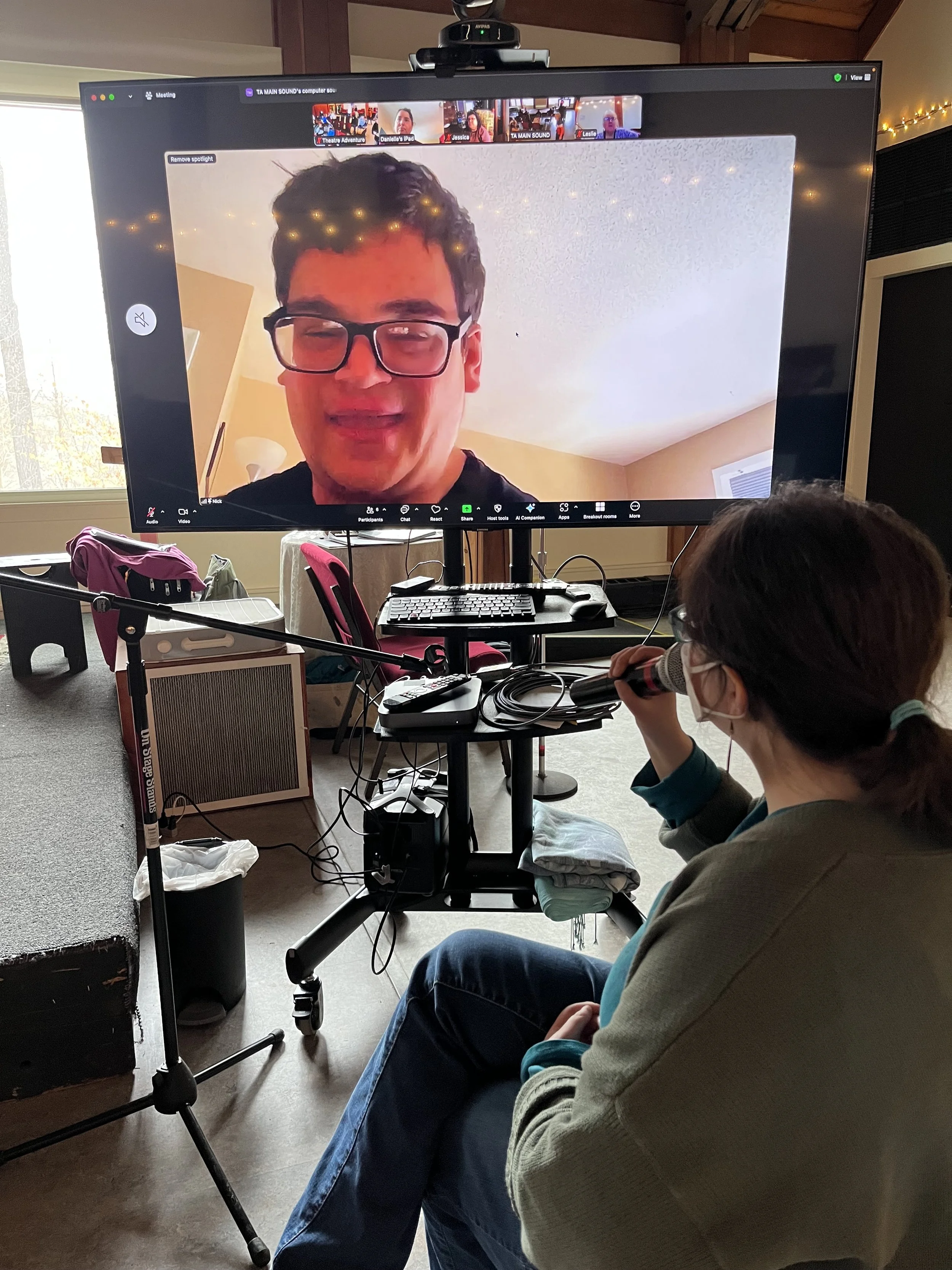 A teacher sits facing a large monitor speaking into a microphone and conversing with an online Troupe Member featured in their zoom window on the monitor.