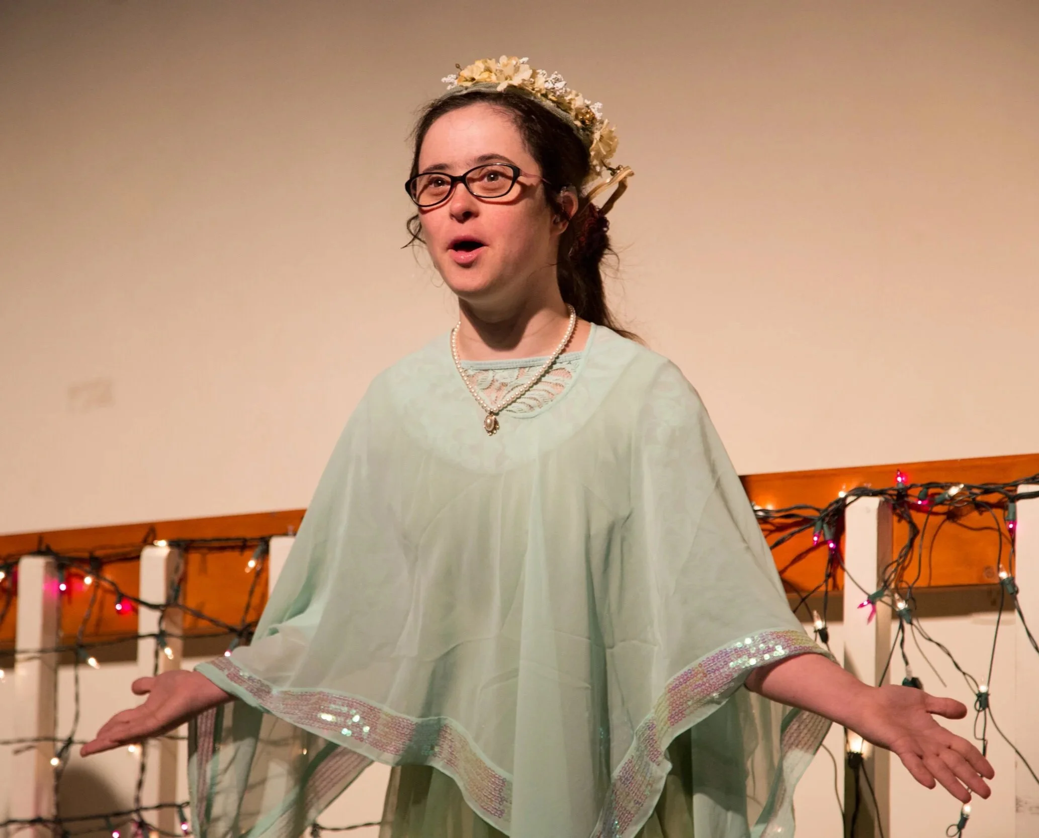 An actor performs wearing a period costume. A sheer mint-colored silky shawl and flowered crown. Her arms and hands are opening out from her sides.