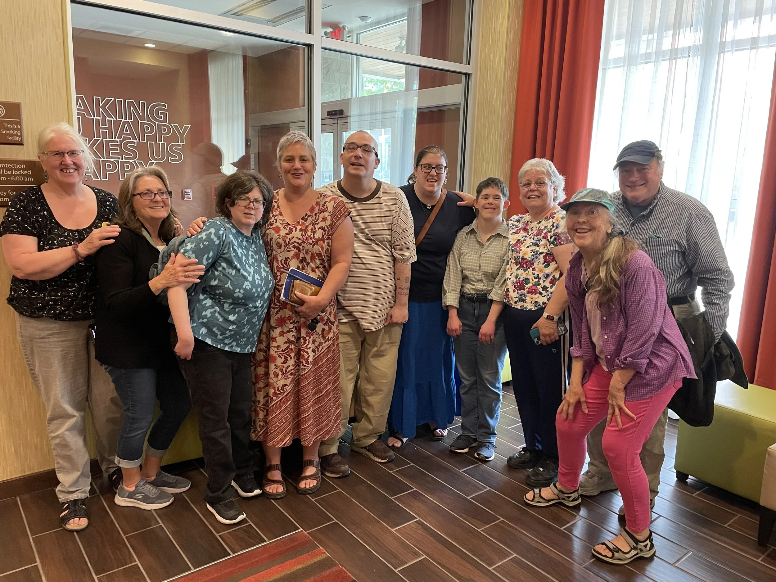 Teachers, friends, and Traveling Troupe members stand shoulder to shoulder, smiling for a group photo in a well-lit lobby.