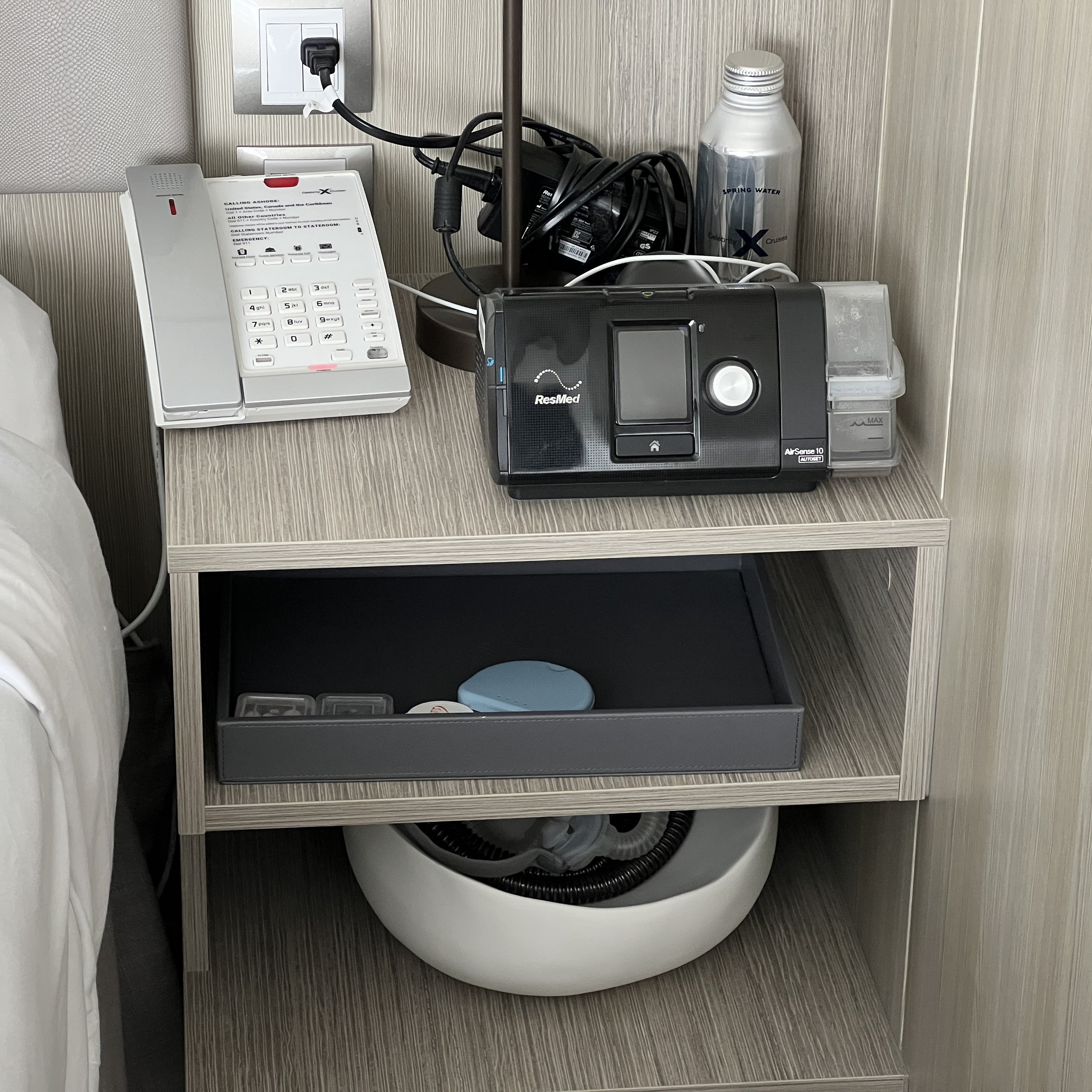 A wider nighstand on the right side of the bed. On top is the phone and a CPAP machine owned by the cruiser. Two open shelves are below holding belongings of the cruiser.