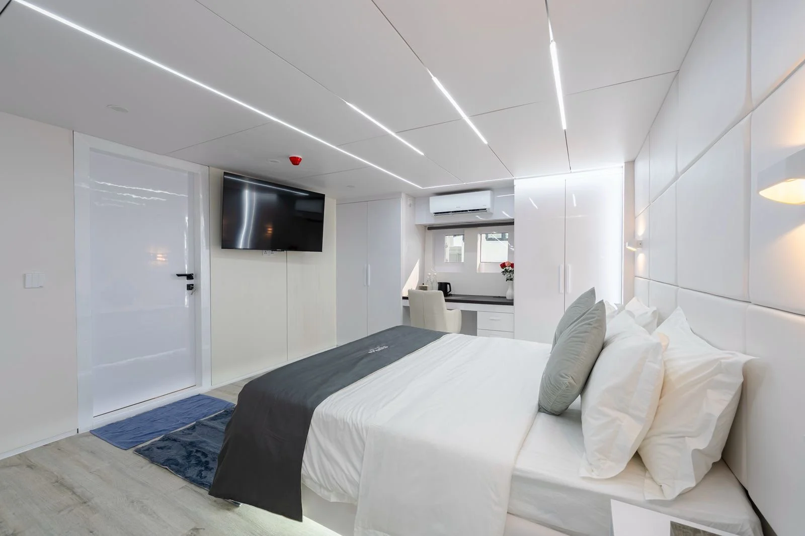 Modern hotel room with white walls, a bed with white linens and gray pillows, a wall-mounted TV, a small desk with a chair, windows with blinds, and a towel rug on the floor.