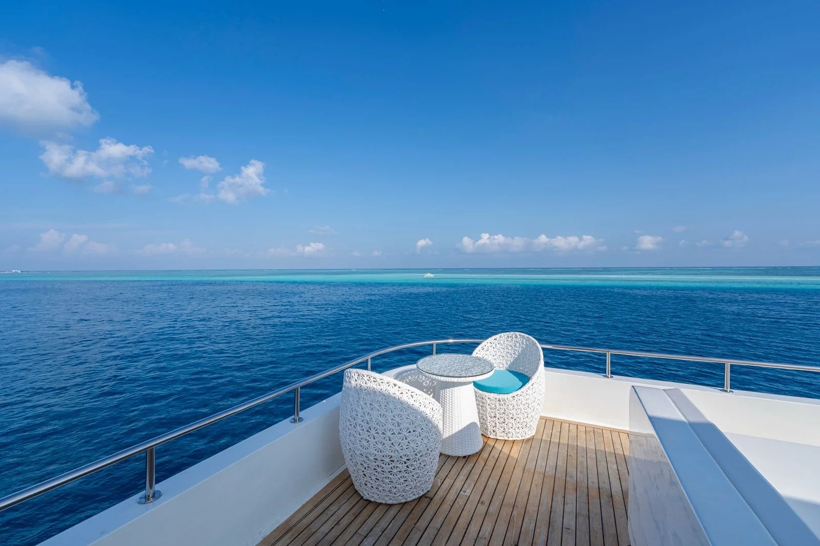 A view from a boat's deck showing the ocean with clear blue water, a distant boat, and clouds in a bright sky. There is a white wicker table with two matching chairs with blue cushions on the deck.