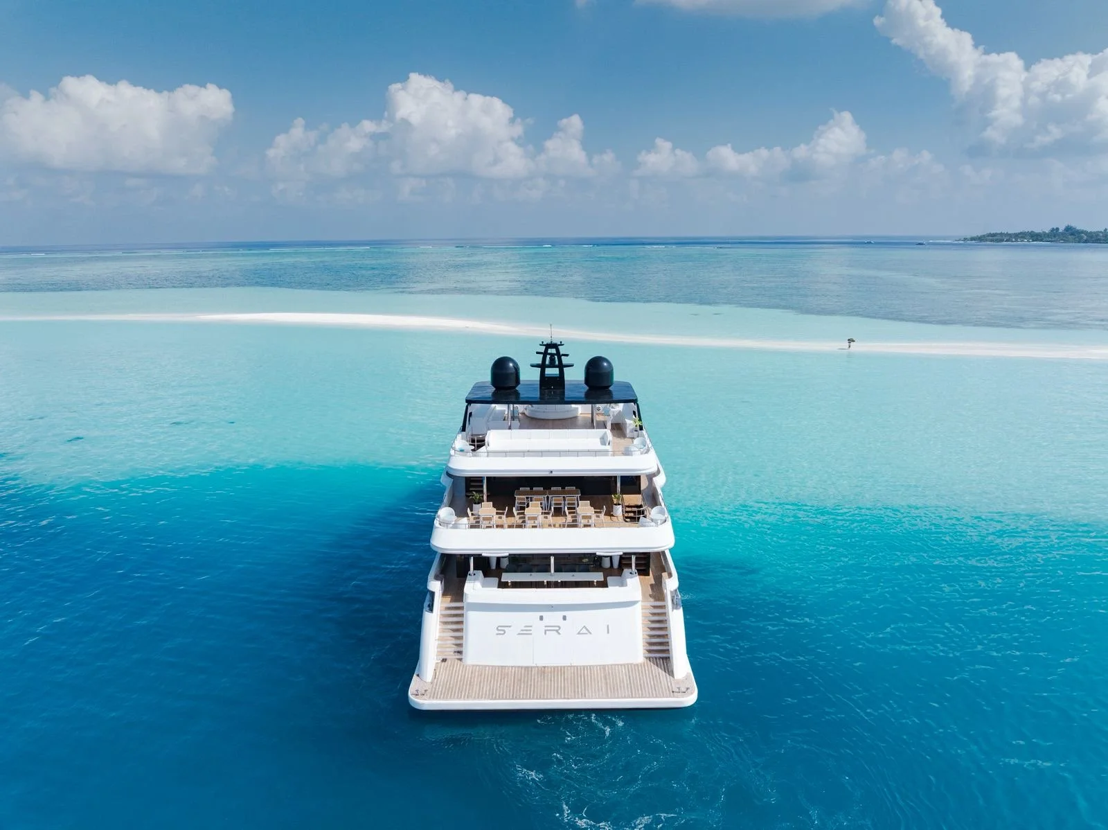A large white luxury yacht sailing on clear blue ocean water with a sandy beach and land visible in the distance under a blue sky with some clouds.