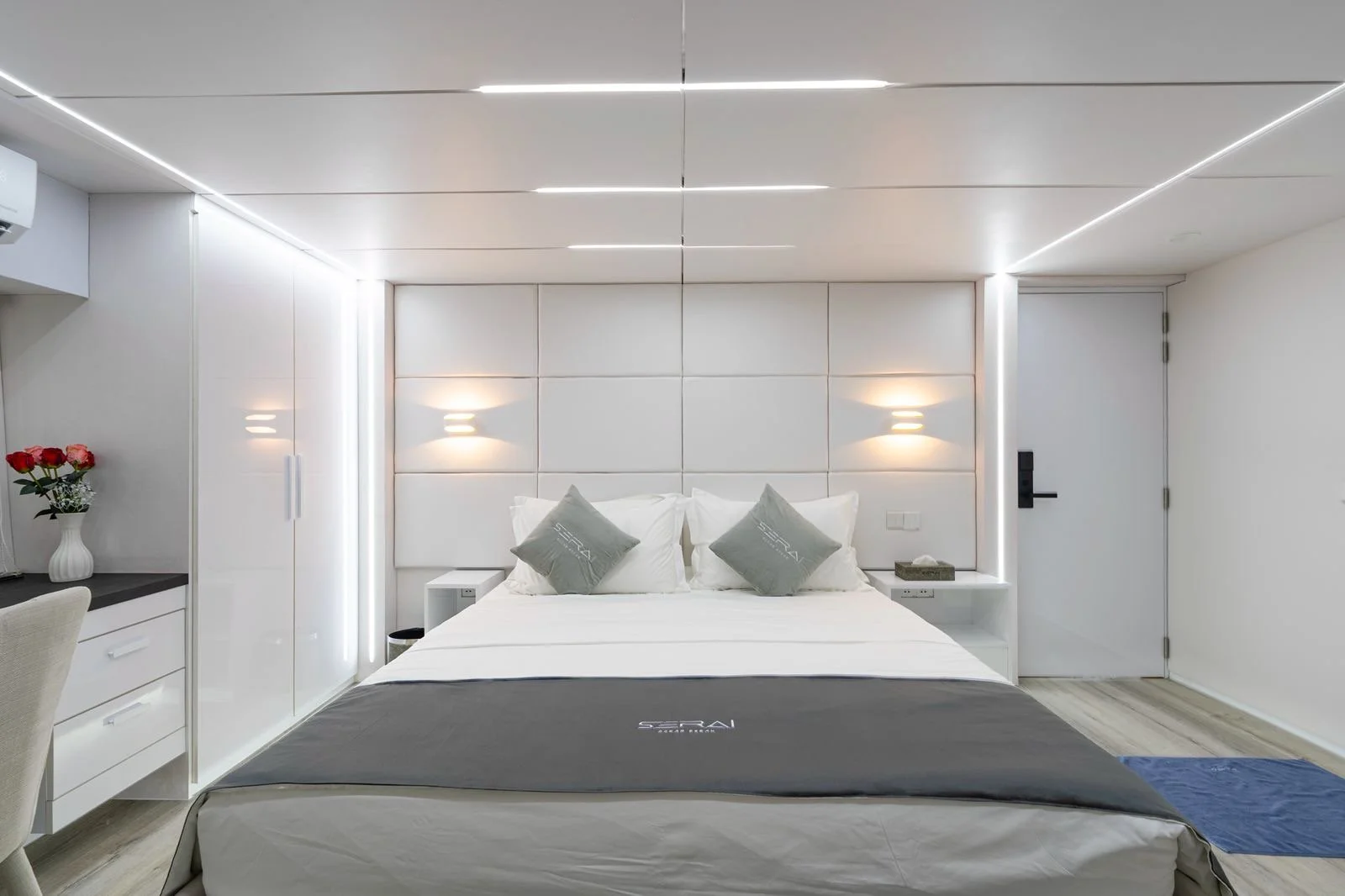 Modern hotel room with a white bed, gray pillows, wall-mounted lights, and a white wardrobe.