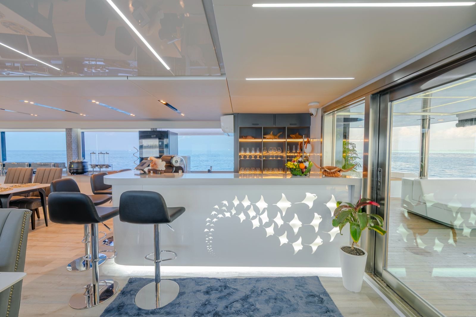 Modern yacht interior with a white bar counter, black bar stools, seating area, ocean view through large windows, and a potted plant near a sliding glass door