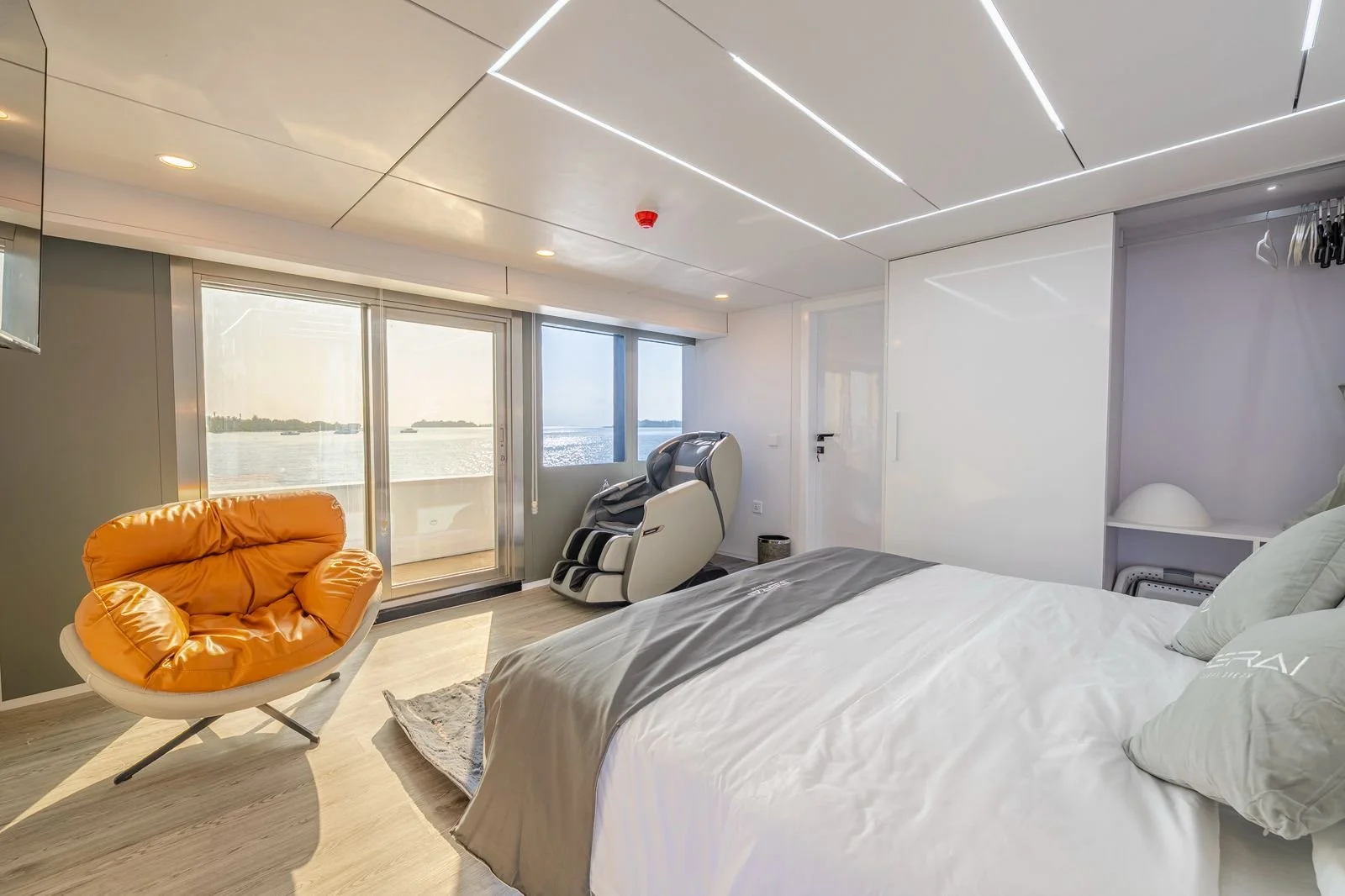 Modern bedroom with large window overlooking water, featuring an orange lounge chair, a massage chair, and a bed with white linens.