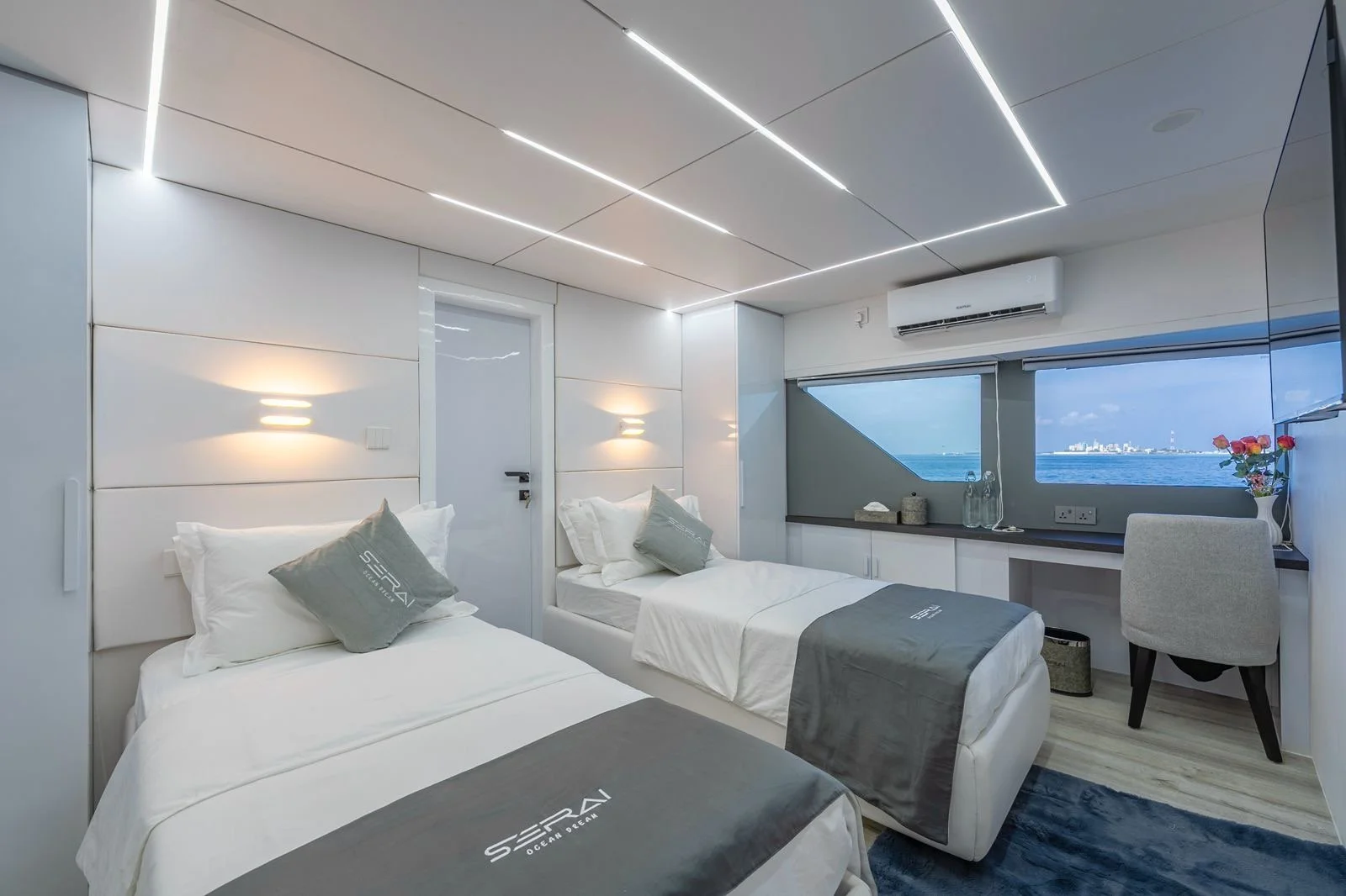 Hotel room with two white beds, gray bed runners, modern lighting, window showing ocean view, air conditioner, desk with flowers, and light-colored decor.