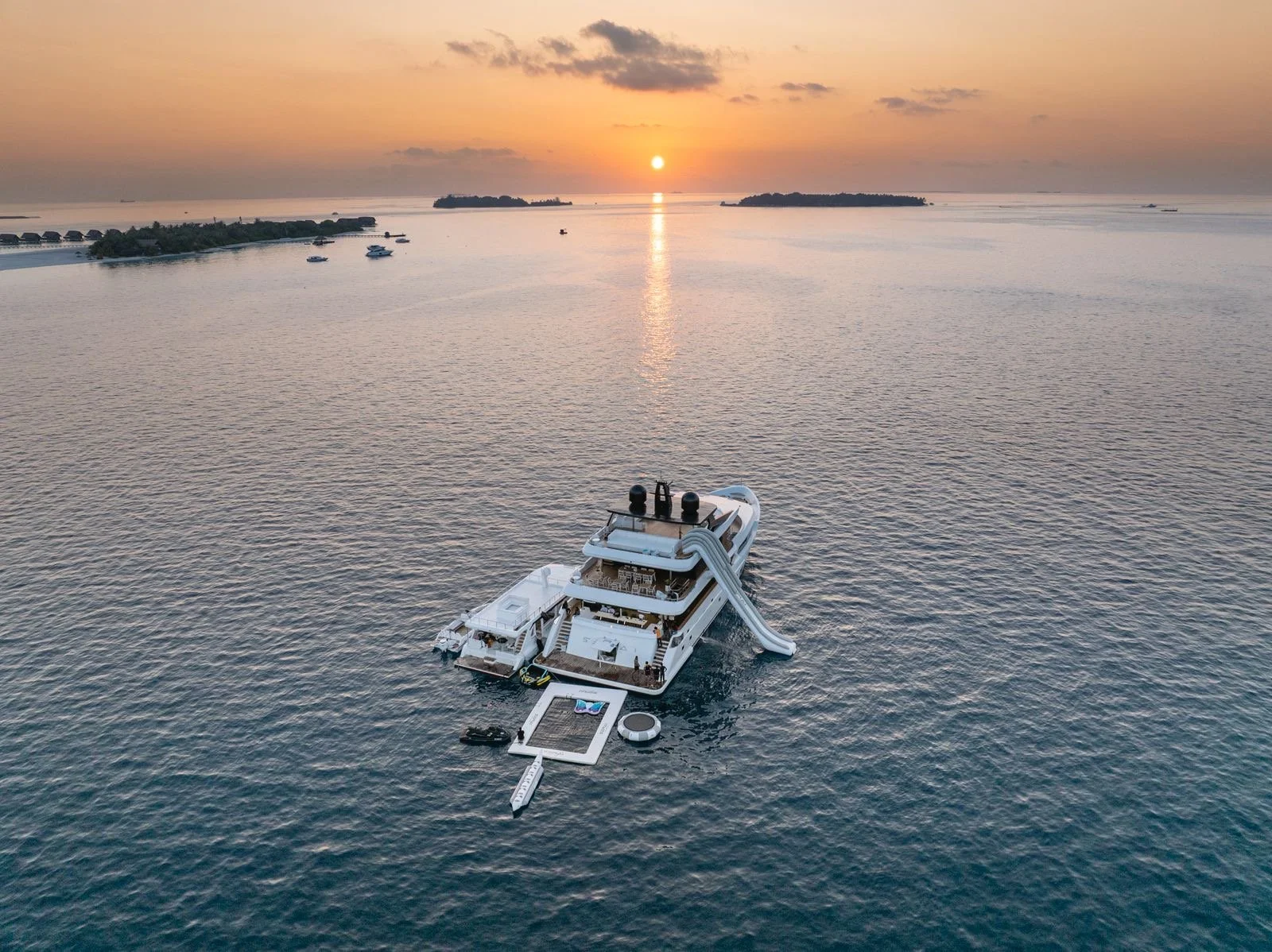 A large yacht with a water slide on its side, anchored in calm ocean water during sunset.