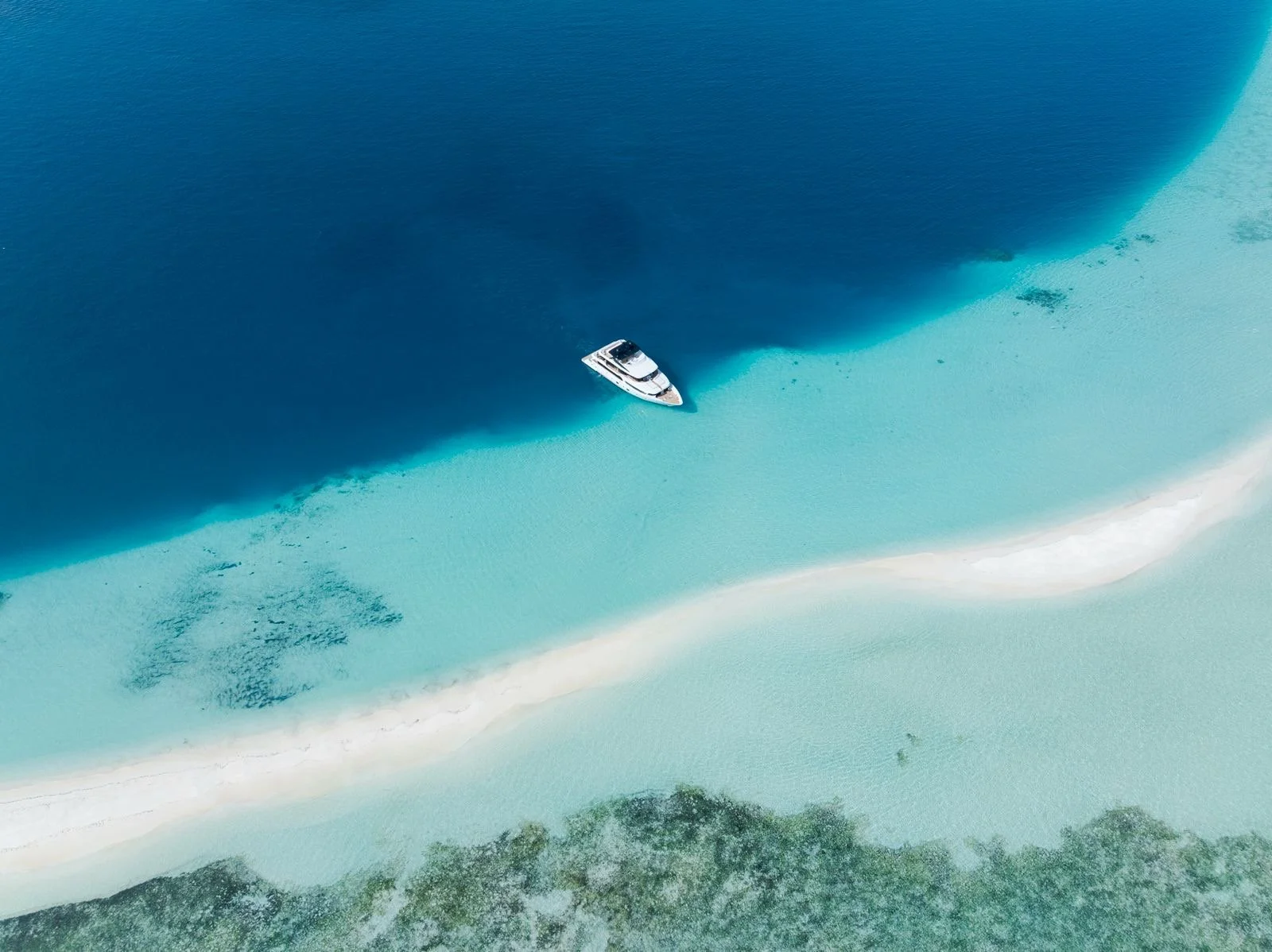 An aerial view of a yacht anchored in turquoise waters near a strip of white sand beach, with green land at the bottom of the image.