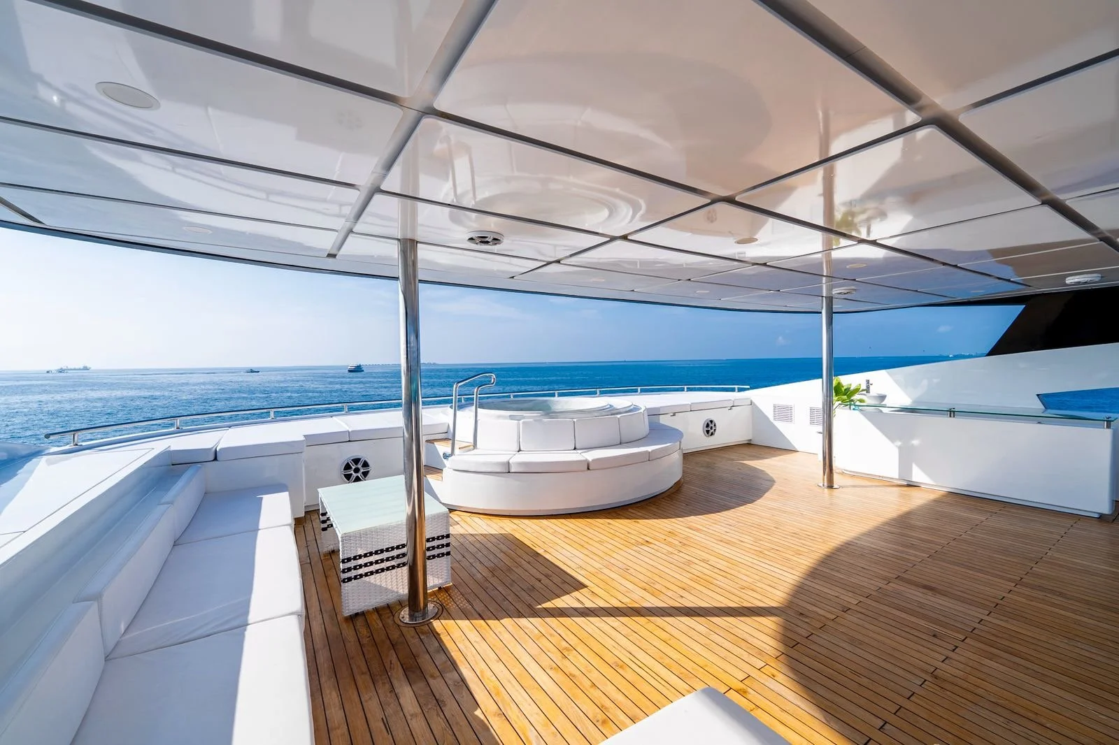 A spacious yacht deck with white cushioned seating along the sides, a small table, a round hot tub, and the ocean in the background under a partly cloudy sky.