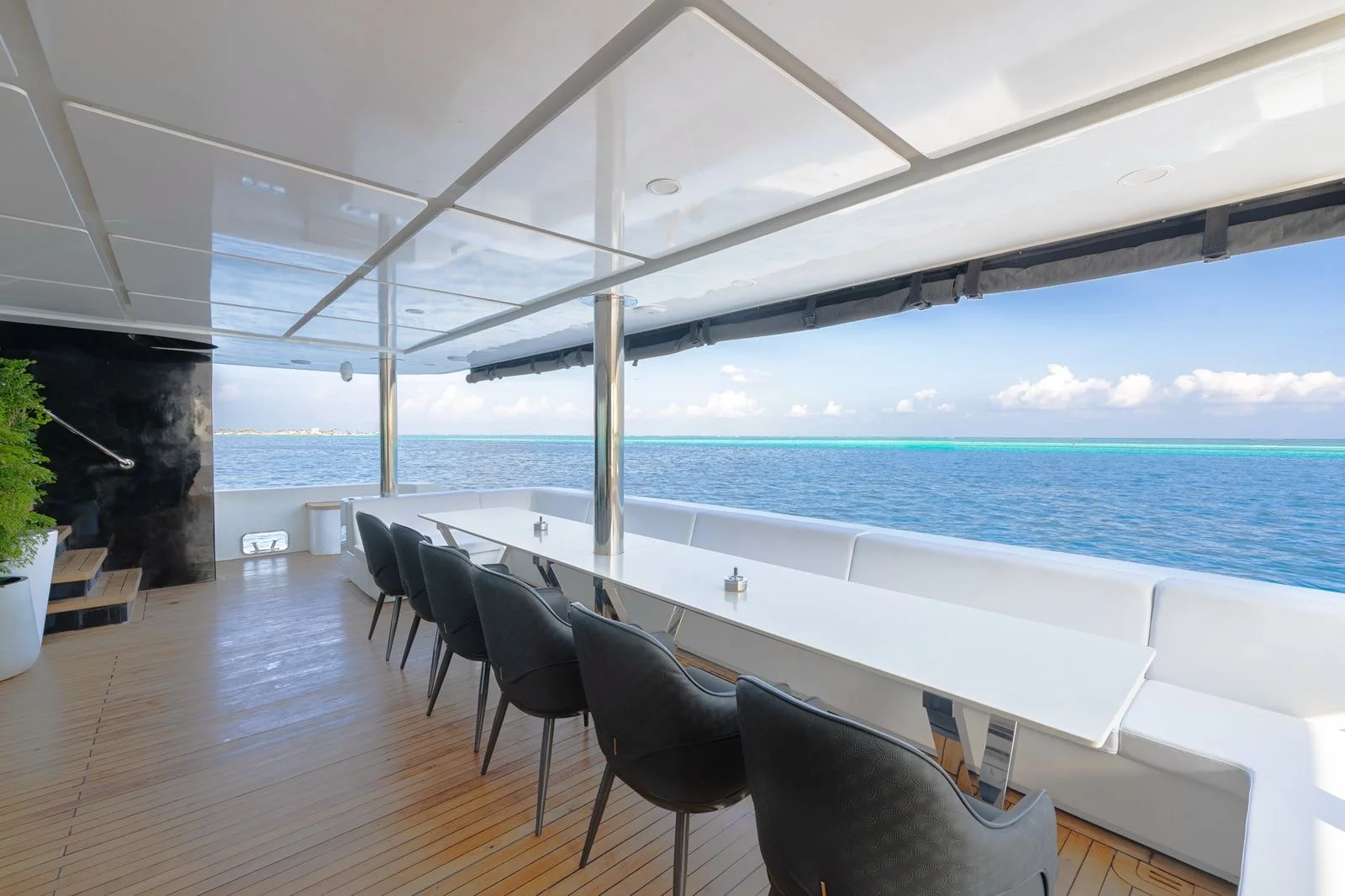 An outdoor dining area on a yacht with a long white table, black chairs, and ocean view with clear blue sky and clouds.