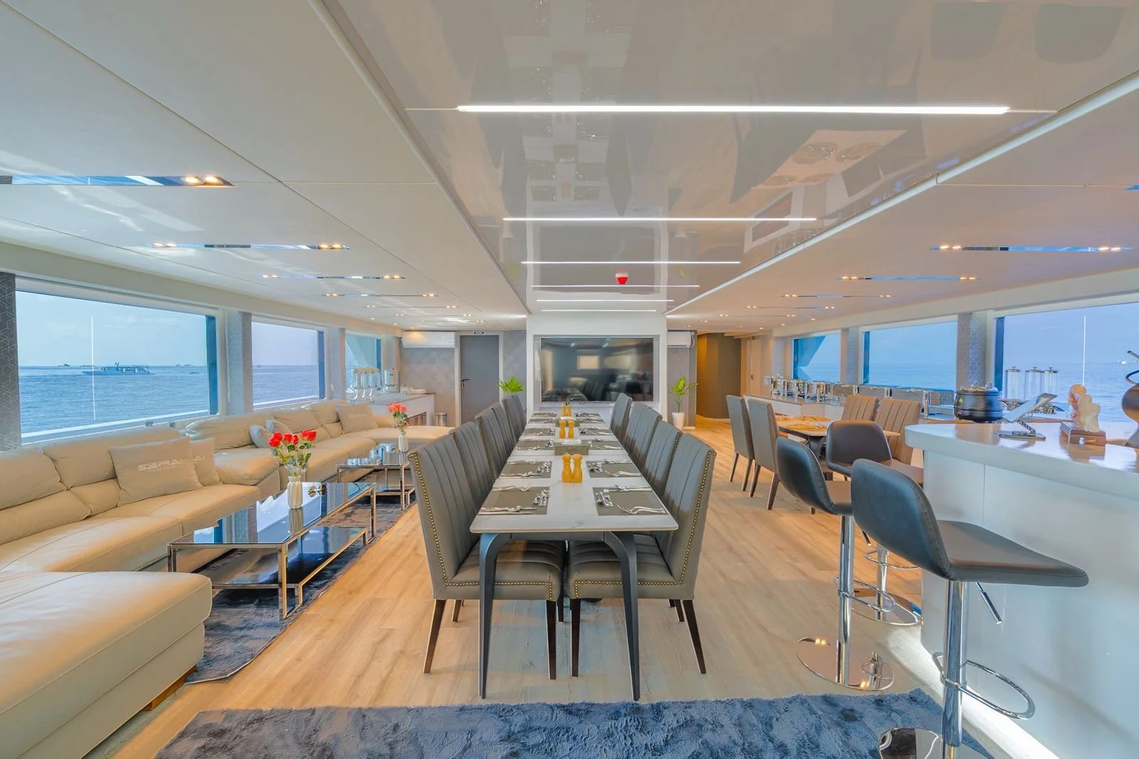 Luxurious dining area on a cruise ship with large windows overlooking the ocean, a long table set for a meal, and a bar counter with stools.