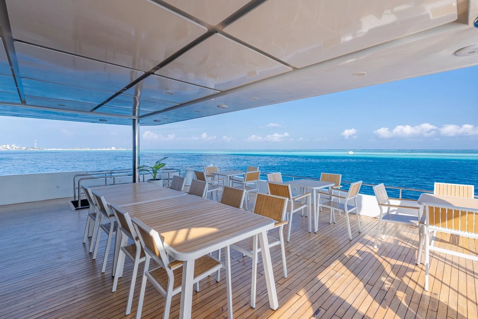 Outdoor deck area on a cruise ship with wooden flooring, tables, and chairs, overlooking the ocean with a city skyline in the distance on a clear day.