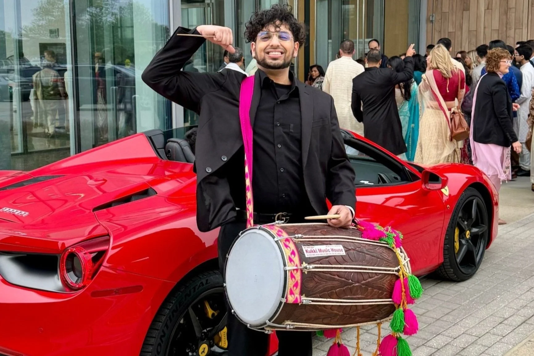 Man in black suit and glasses playing a drum with a group of people gathered outside a building, standing next to a red sports car.