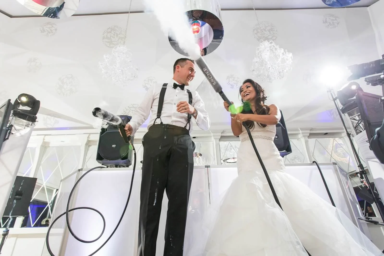 A bride and groom celebrating their wedding with champagne, holding large foam cannons. The bride wears a white wedding gown and the groom in a tuxedo with suspenders. The scene is lively with musical equipment and chandeliers in the background.