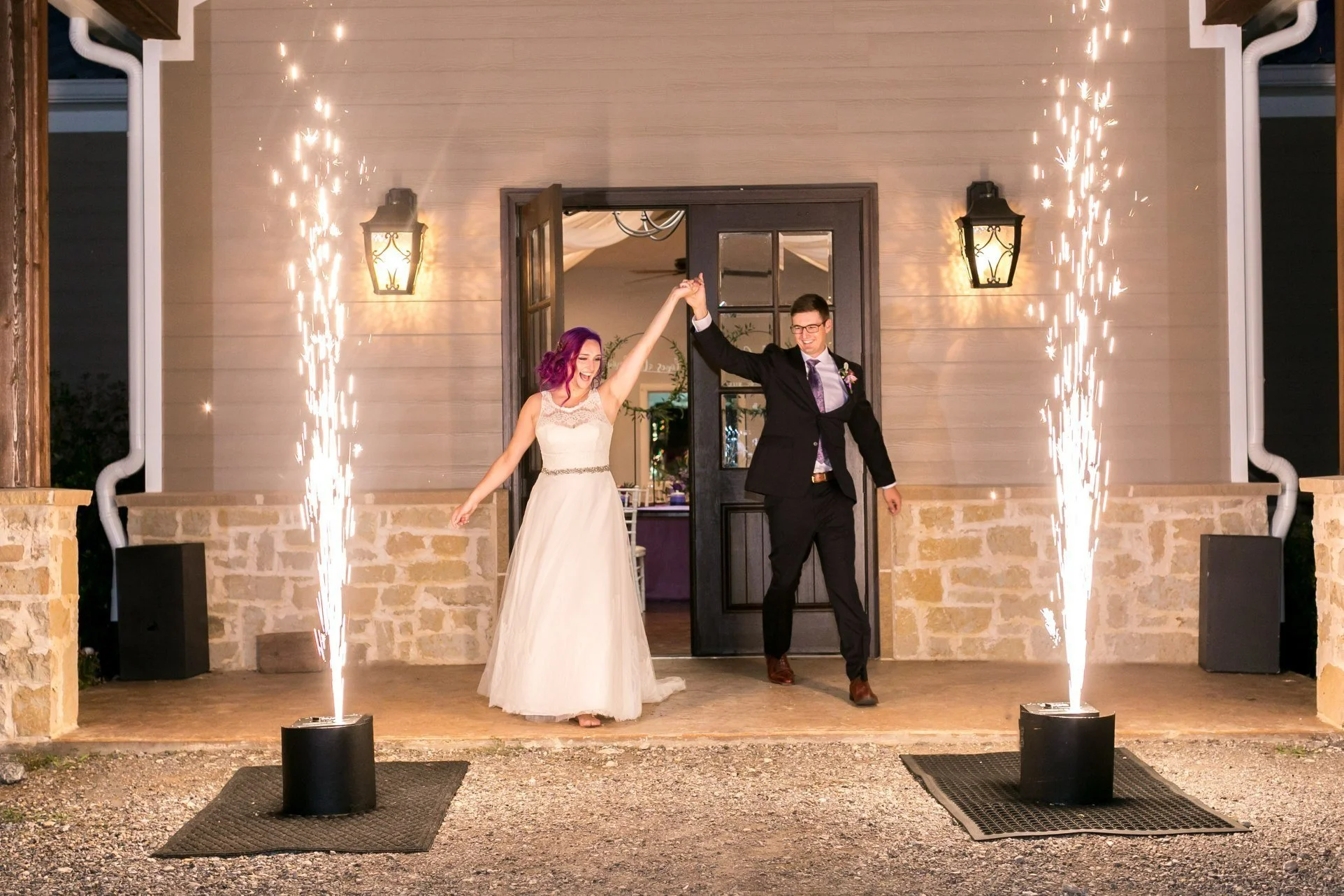 A newlywed couple dancing outside a building with sparklers on either side, one dancer woman in a white dress with purple hair, the male in a black suit and glasses, celebrating under warm outdoor lighting.
