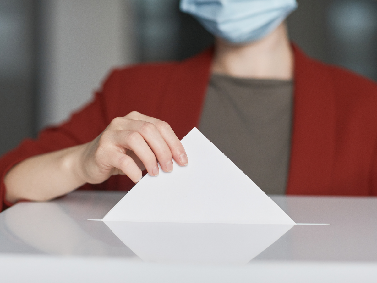 Person wearing a mask and a red blazer placing their ballot into a ballet box