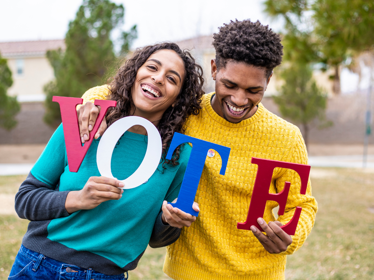 A woman and a man in brightly colored clothing holding up the letters to the word "Vote" with the "V" and "E" in the man's hands that are wrapped around the woman holding the "O" and "T"