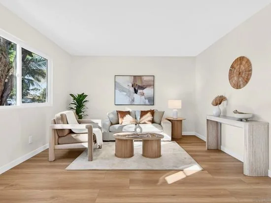 Living room with white walls, wooden floor, two windows with a view of palm trees, a white sofa with decorative pillows, a beige armchair, a round coffee table, a potted plant, a side table with a lamp, and decorative wall art.