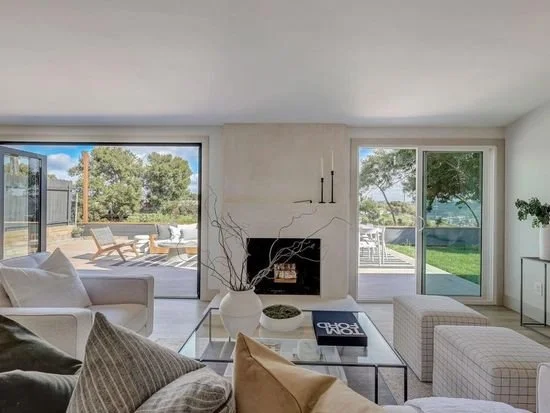 Modern living room with large glass doors leading to outdoor patio and garden, white sofas, and a fireplace.