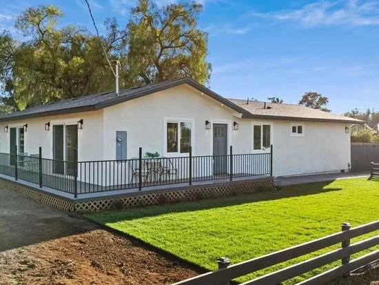 Single-story white house with a black deck railing surrounded by a green lawn and trees under a blue sky.