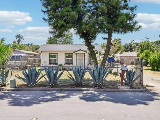 Single-story house with a gray roof surrounded by agave plants and a large tree, fenced yard, suburban neighborhood with blue sky and clouds.