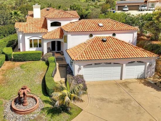 A white house with a red-tiled roof, two-car garage, surrounded by a well-maintained lawn and landscaped garden, including a fountain and palm tree.