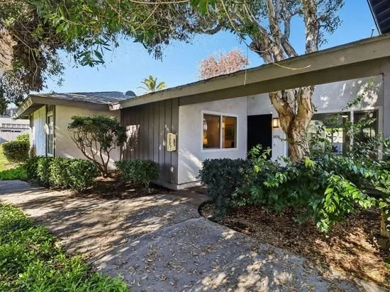 A small residential house with gray and white exterior, surrounded by trees and bushes, with a concrete walkway leading to the entrance.