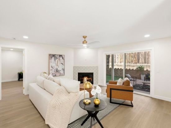 Bright living room with white walls and a fireplace, beige sofa, wooden armchair, glass sliding doors, and outdoor patio visible.