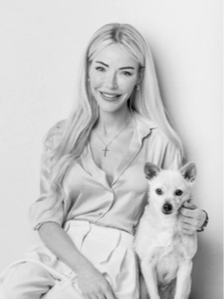 A young woman with long blonde hair smiling and sitting next to a small dog, both facing the camera in a black-and-white photo.