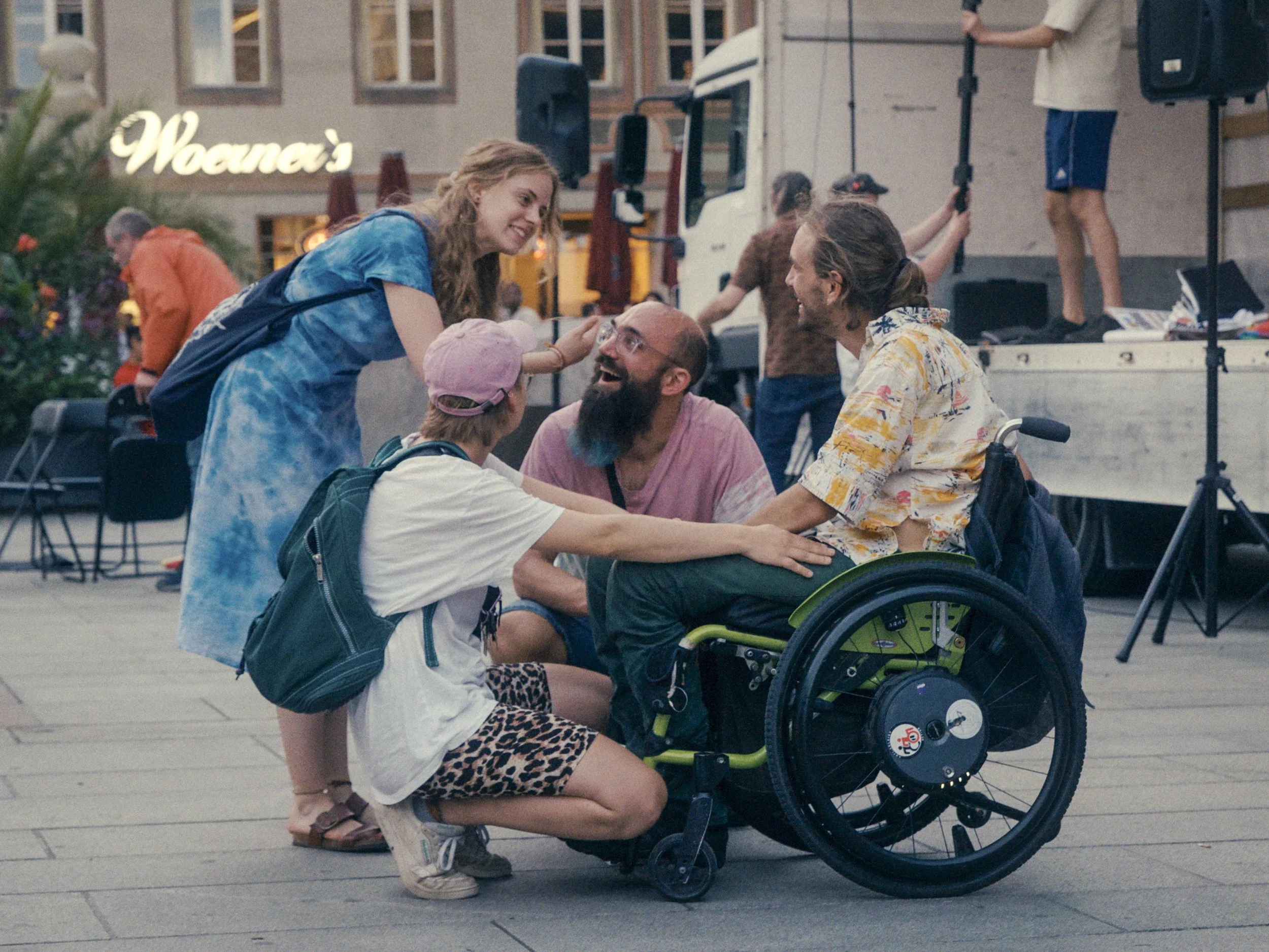 Vier Personen auf einem Platz. Eine Im Rollstuhl. Die anderen in der Hocke um diese Person herum.