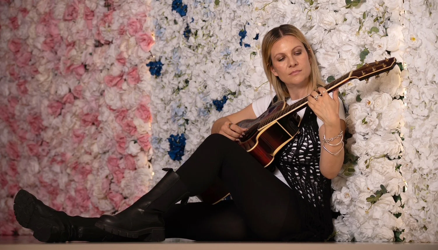 Sabine die Workshopleiterin lehnt sitzend an einer Blumenwand mit ihrer Gitarre.