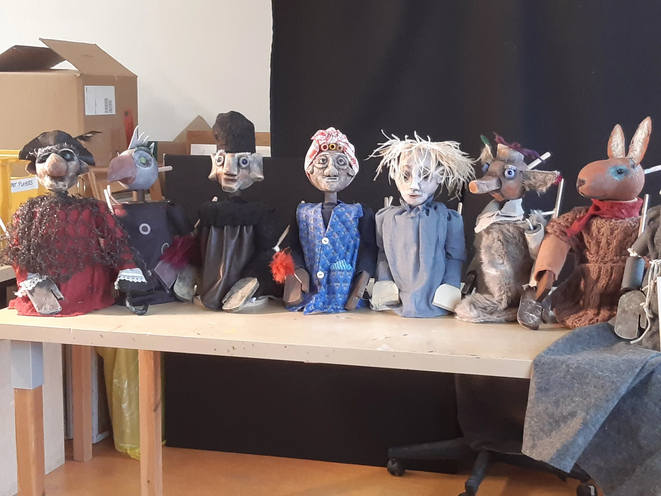 Viele Marionettenfiguren stehen auf einem Tisch, dahinter Kartons und ein Vorhang