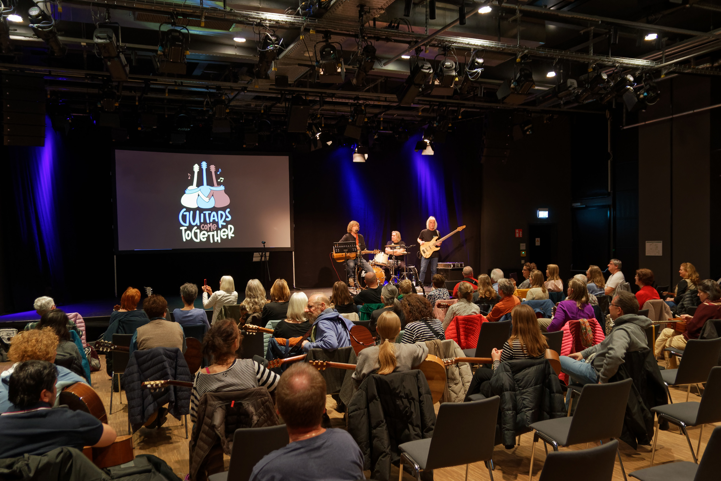 Konzertsaal mit Menschen im Publikum und drei Gitarrenspielern auf de rBühne