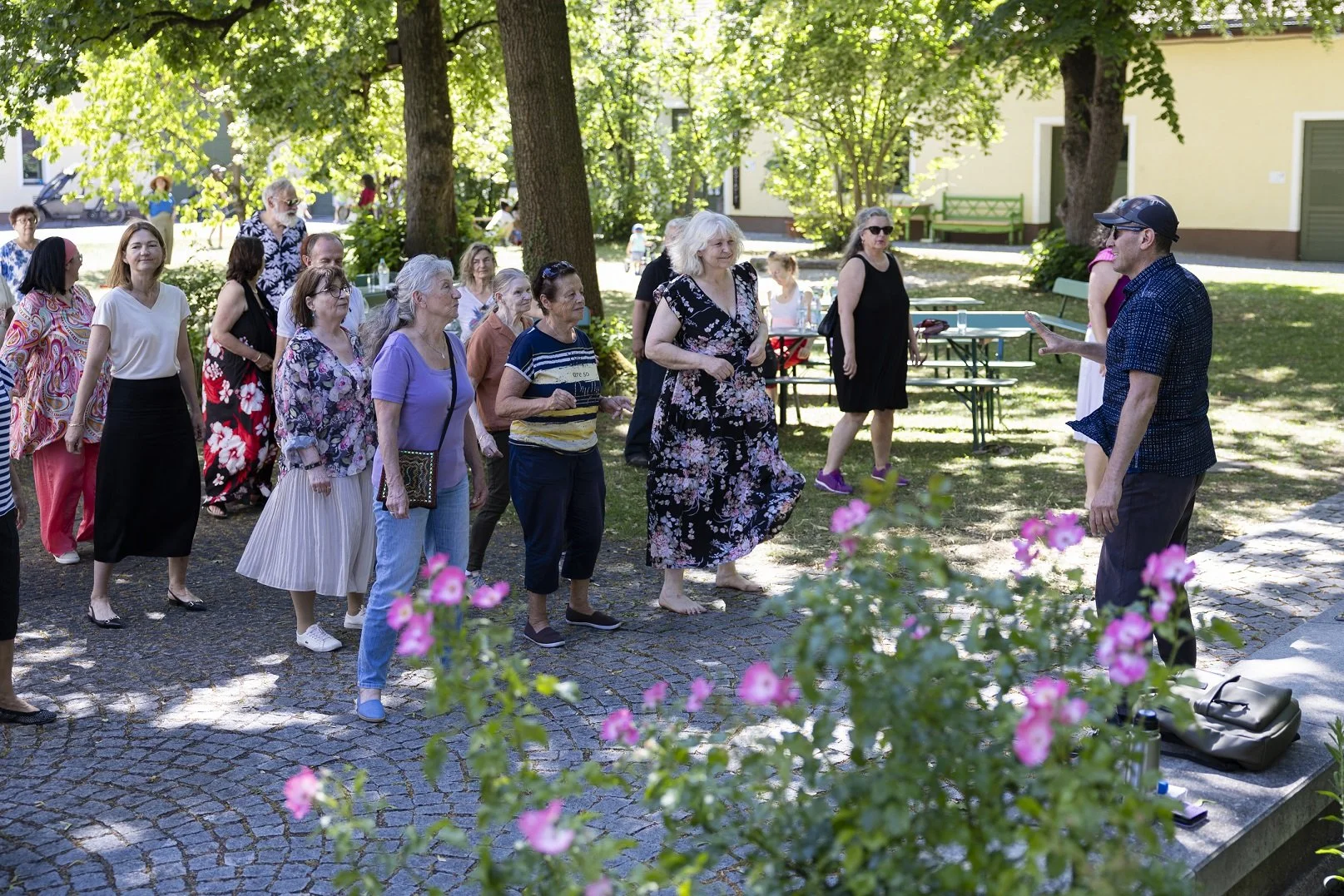 mehreren Menschen draußen unter Bäumen. Sie tanzen. Es gibt Blumen, Bäume und ein paar Tische und Bänke.