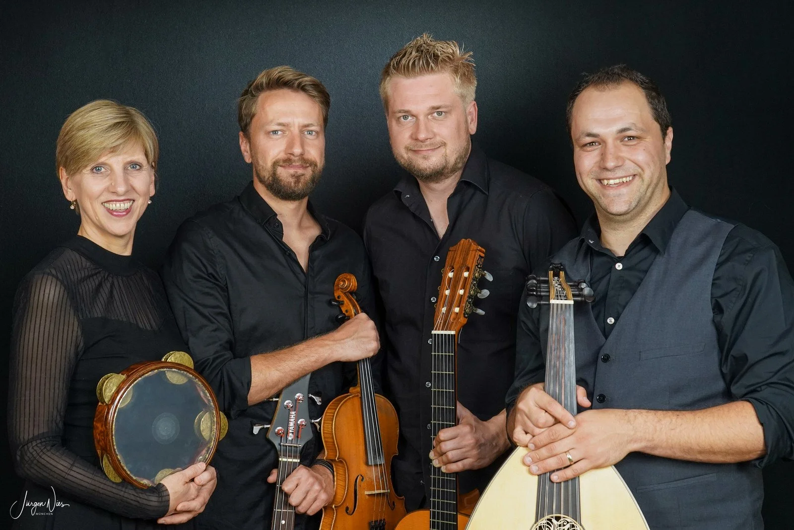 Ein Foto mit 4 Personen vor schwarzem Hintergrund. Sie halten jeweils eine Oud, eine Gitarre, eine Geige und eine Trommel und schauen lächelnd in die Kamera