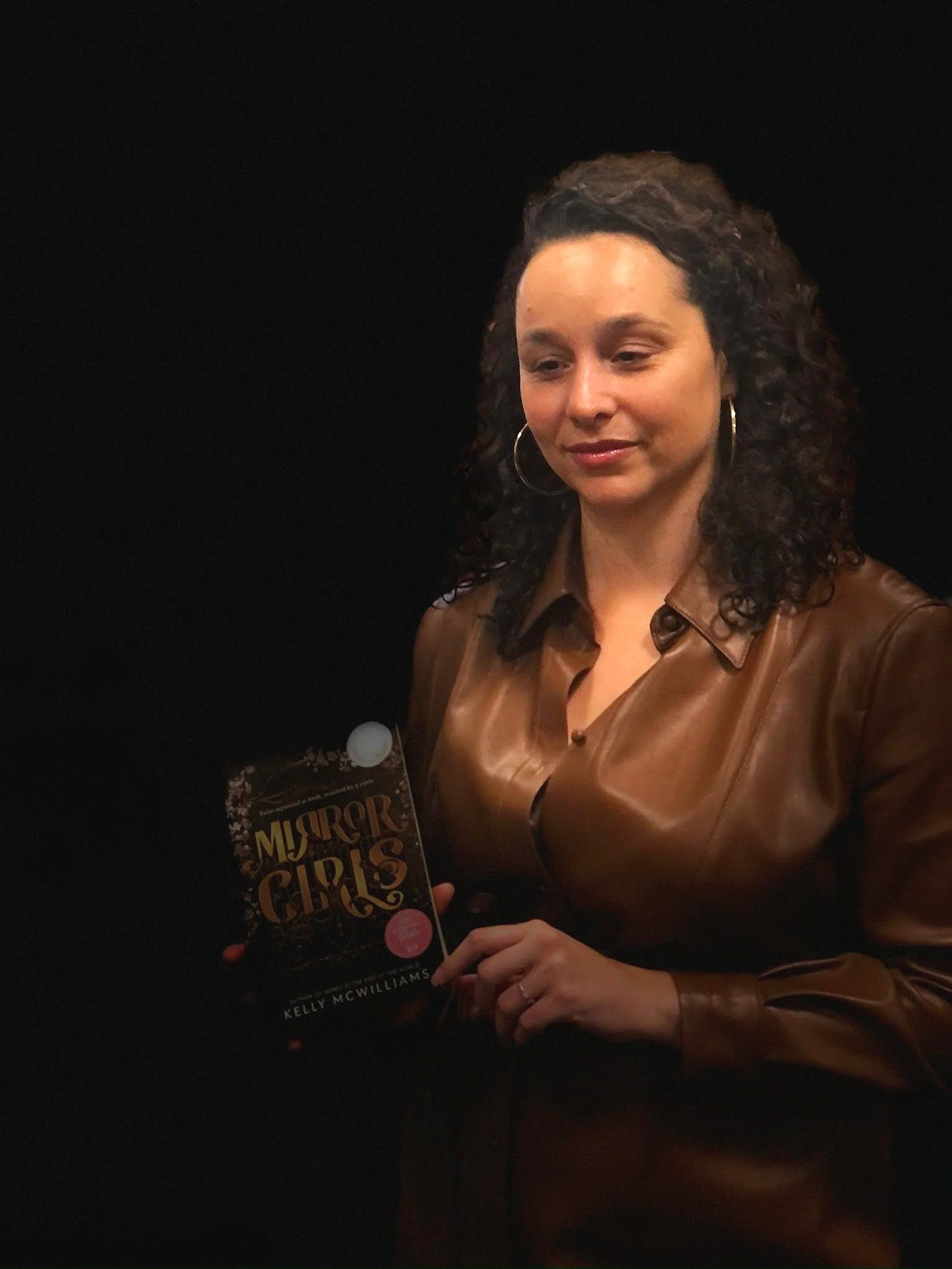 A woman with curly dark hair, wearing hoop earrings and a brown leather jacket, is holding a book titled 'Murder of Girls' by Kelly McWilliams against a dark background.