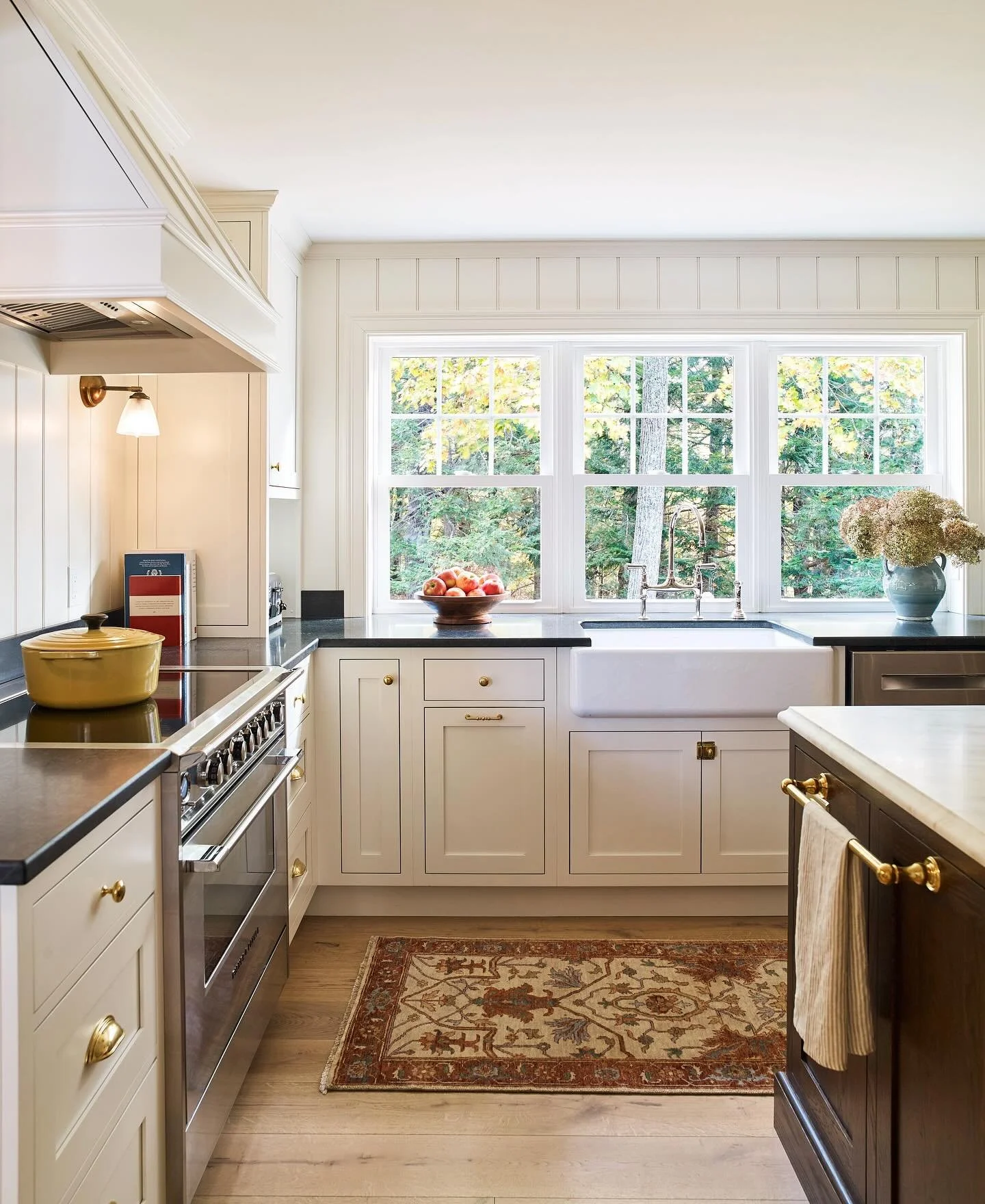 Framed by windows, this Kitchen leans into simplicity with warm white cabinetry, brass hardware and perfectly placed farmhouse sink. The contrast of dark soapstone countertops and light finishes adds depth, while subtle textures - from the runner to 