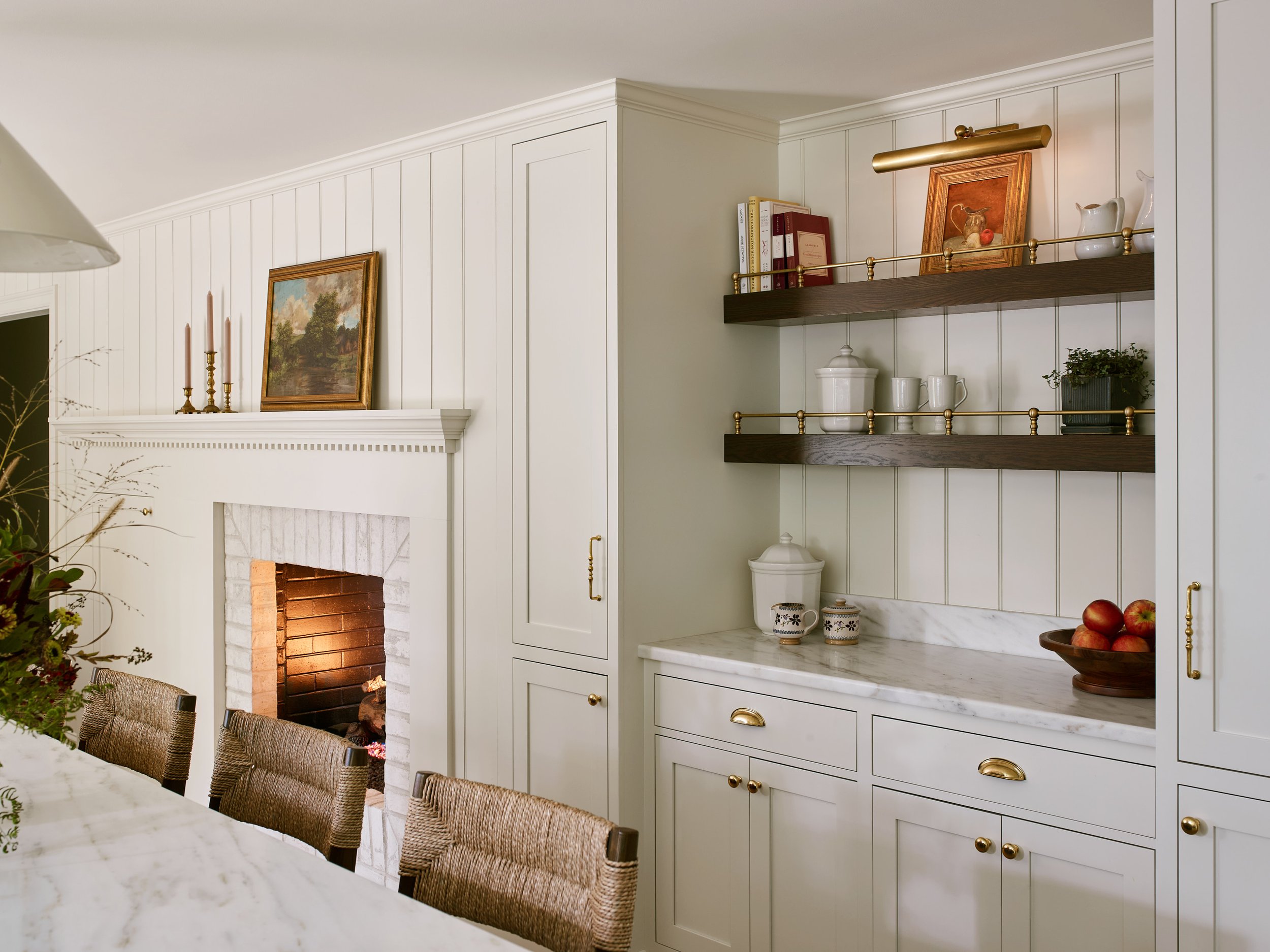 An English kitchen with a white tiled fireplace and mantel, decorated with framed artwork, candles, and a gold candleholder. The kitchen has white cabinets, open wooden shelves with a brass gallery rail, and a Danby marble countertop.