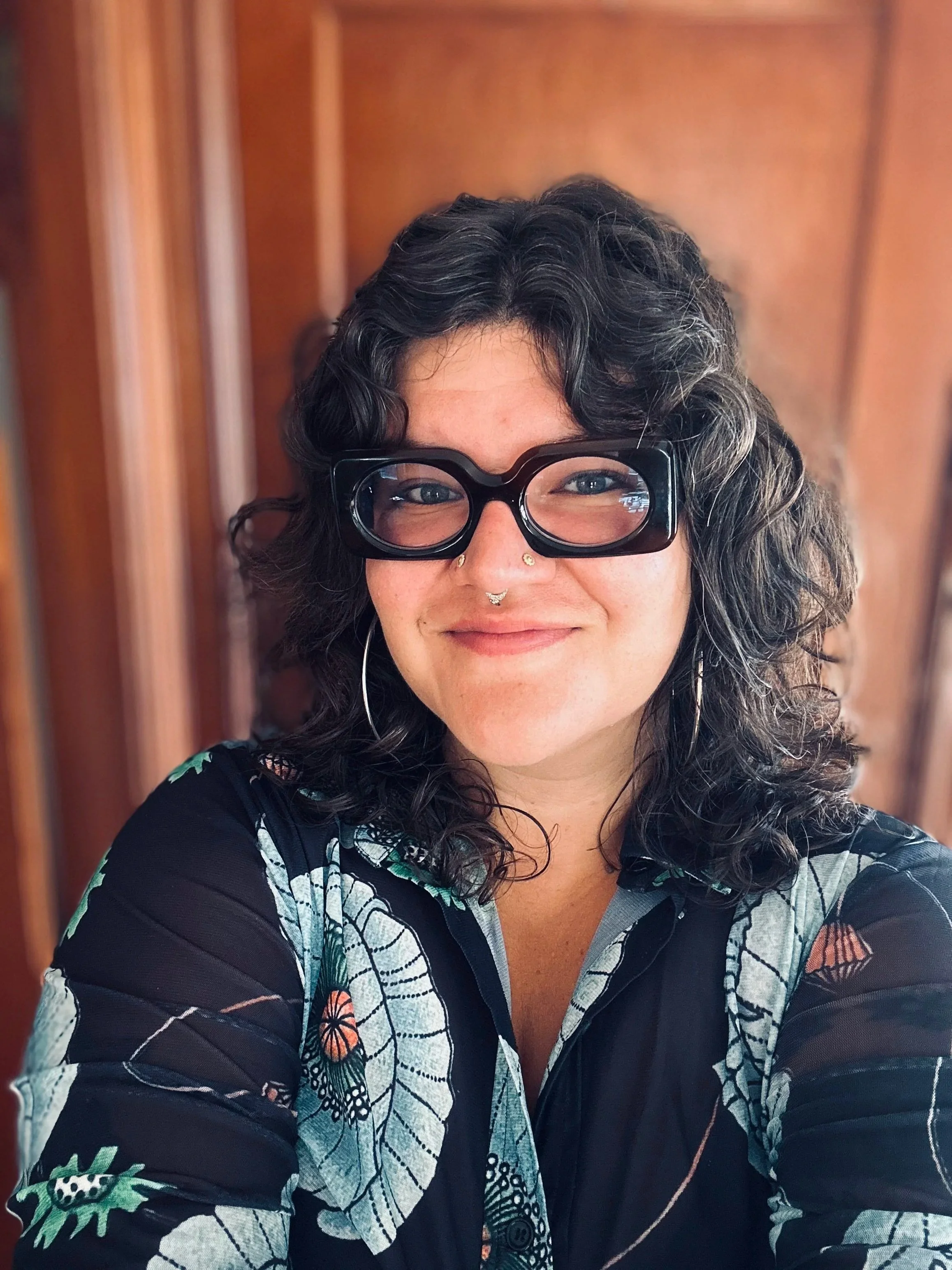 Ali Denis is a queer femme with a cool look. This headshot is from the mid-torso up. Ali wears stylish glasses and has three nose piercings. They have brown wavy hair and are smiling. They wear hoop earrings and a patterned shirt.