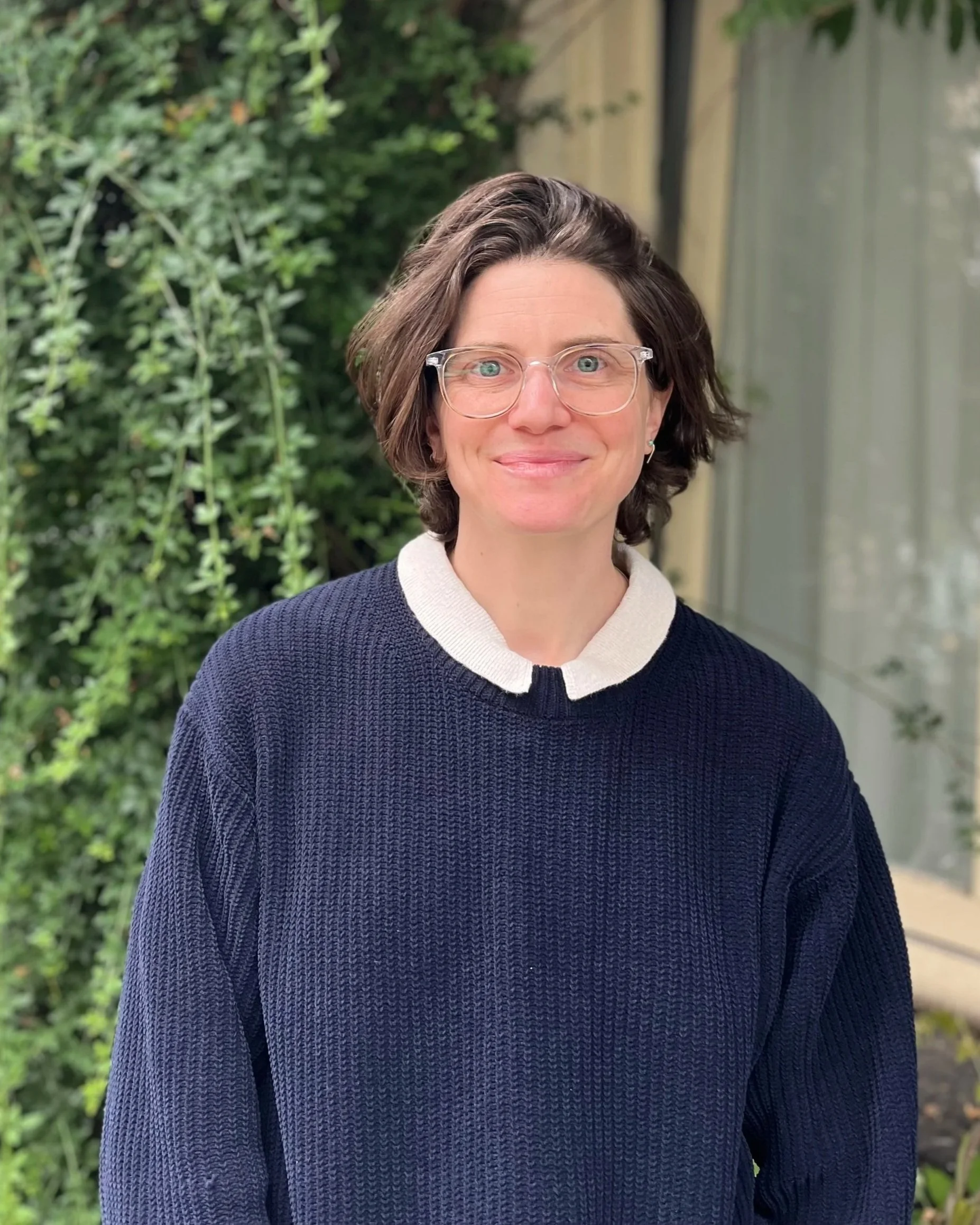 Michele is a white person, standing in nature, looking at the camera with a warm smile. Michele has short brown hair, bright blue eyes, and clear frame glasses. Michelle is wearing a blue sweater with a white collar.