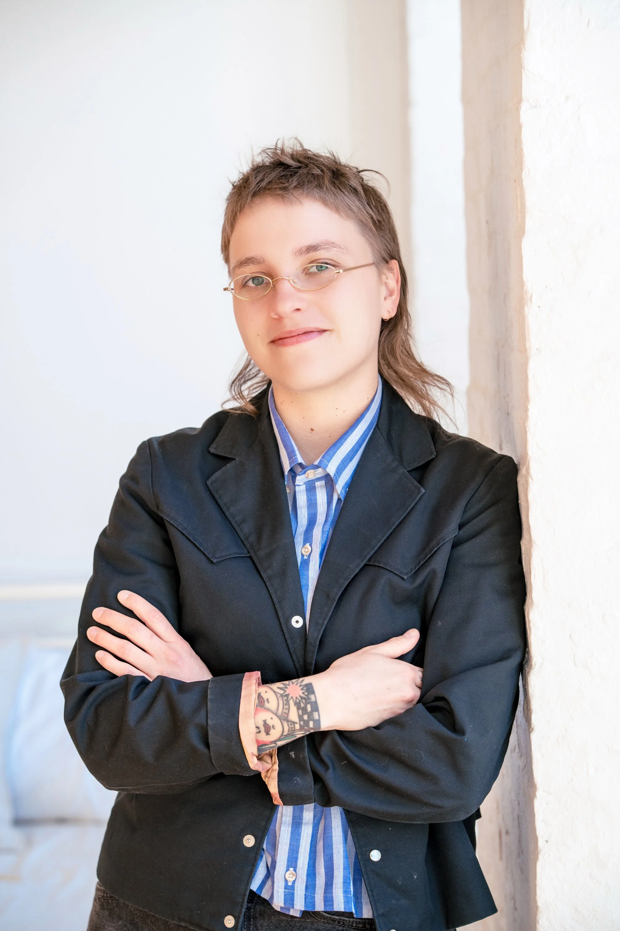Photo of Holly Herzfeld, taken indoors with a neutral background. They are a white adult, has short dark blond hair, blue eyes, wears glasses and has a closed mouth smile. They wear a button down shirt and jacket, and tattoos are visible on their arm