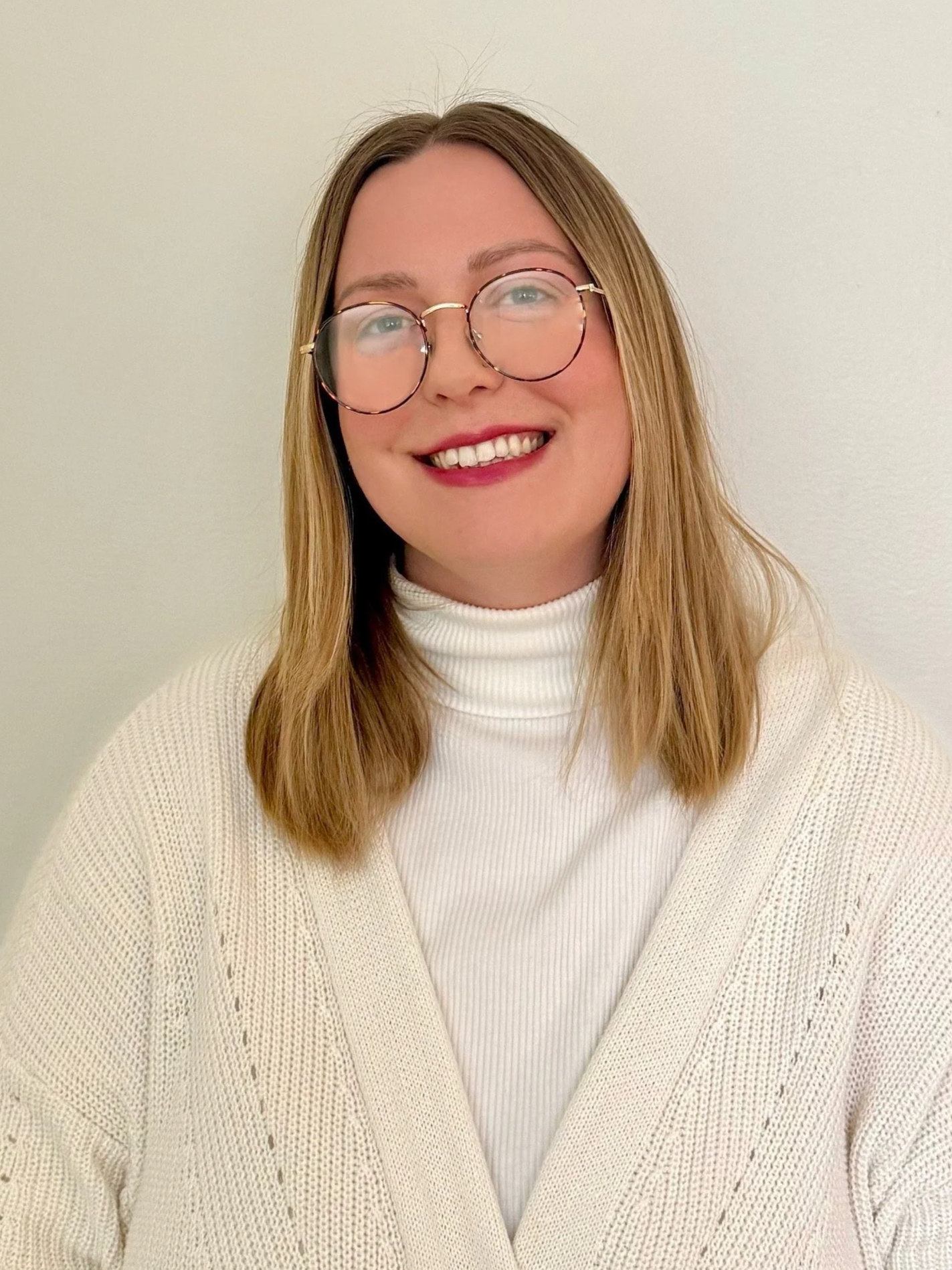 Abby is smiling at the camera and wears red lipstick. She has mid-length blonde hair and wears round tortoise-shell glasses. She is is wearing a white turtleneck and a cream cardigan.