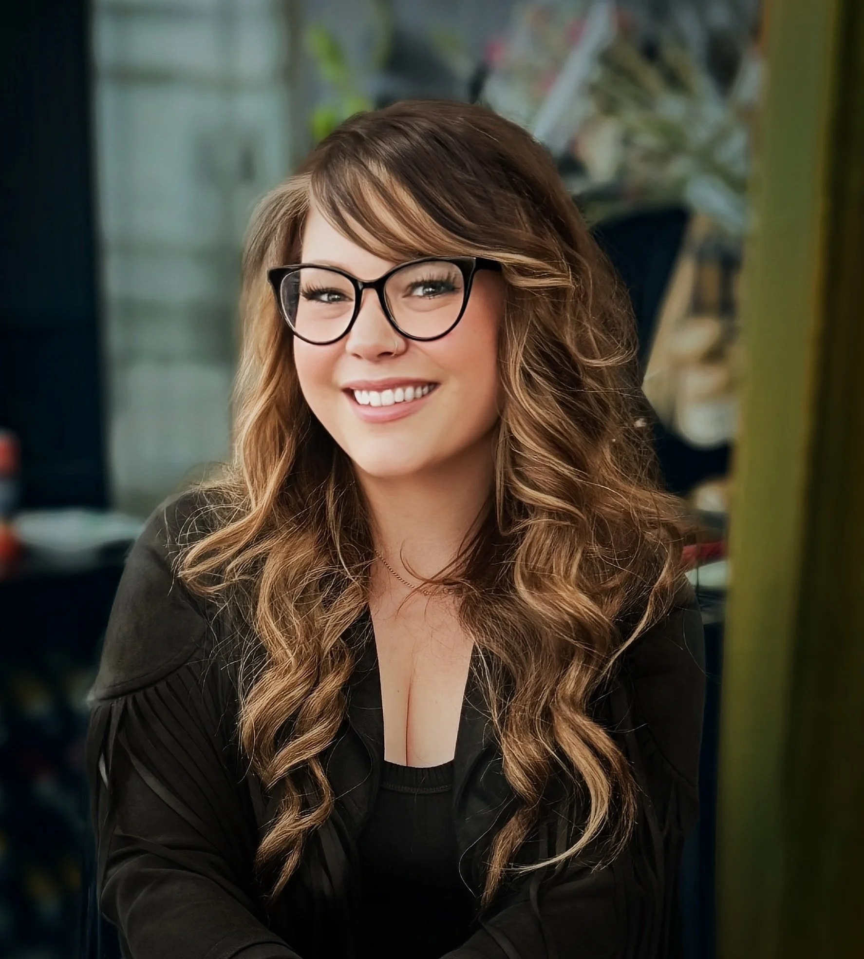 A woman with long wavy brown hair, wearing glasses and a nose ring, smiling, in an indoor setting with a blurred background.
