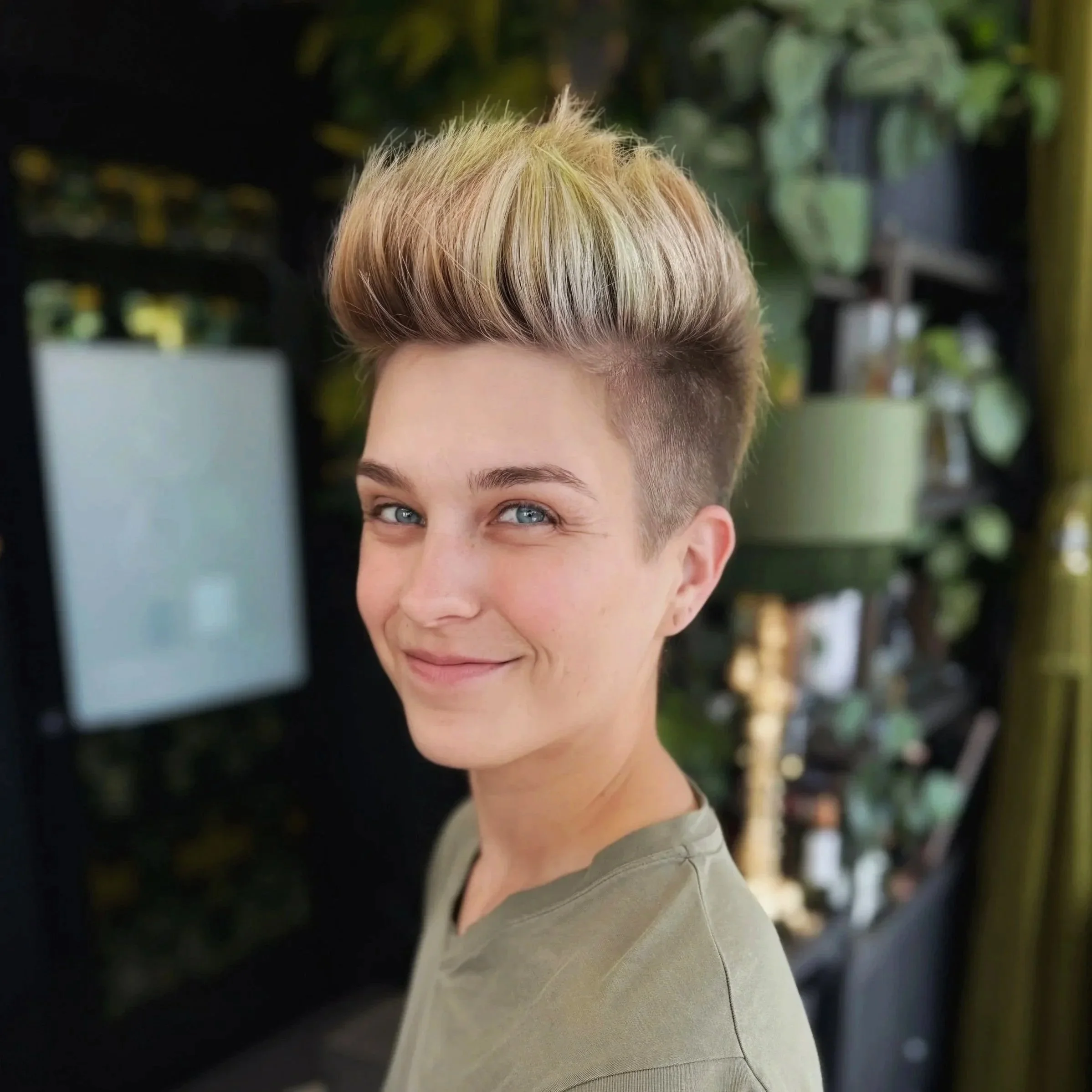 Close-up of a smiling woman with short, blonde hair styled in a spiky updo, wearing a gray top, outdoors with blurred greenery and structures in the background.